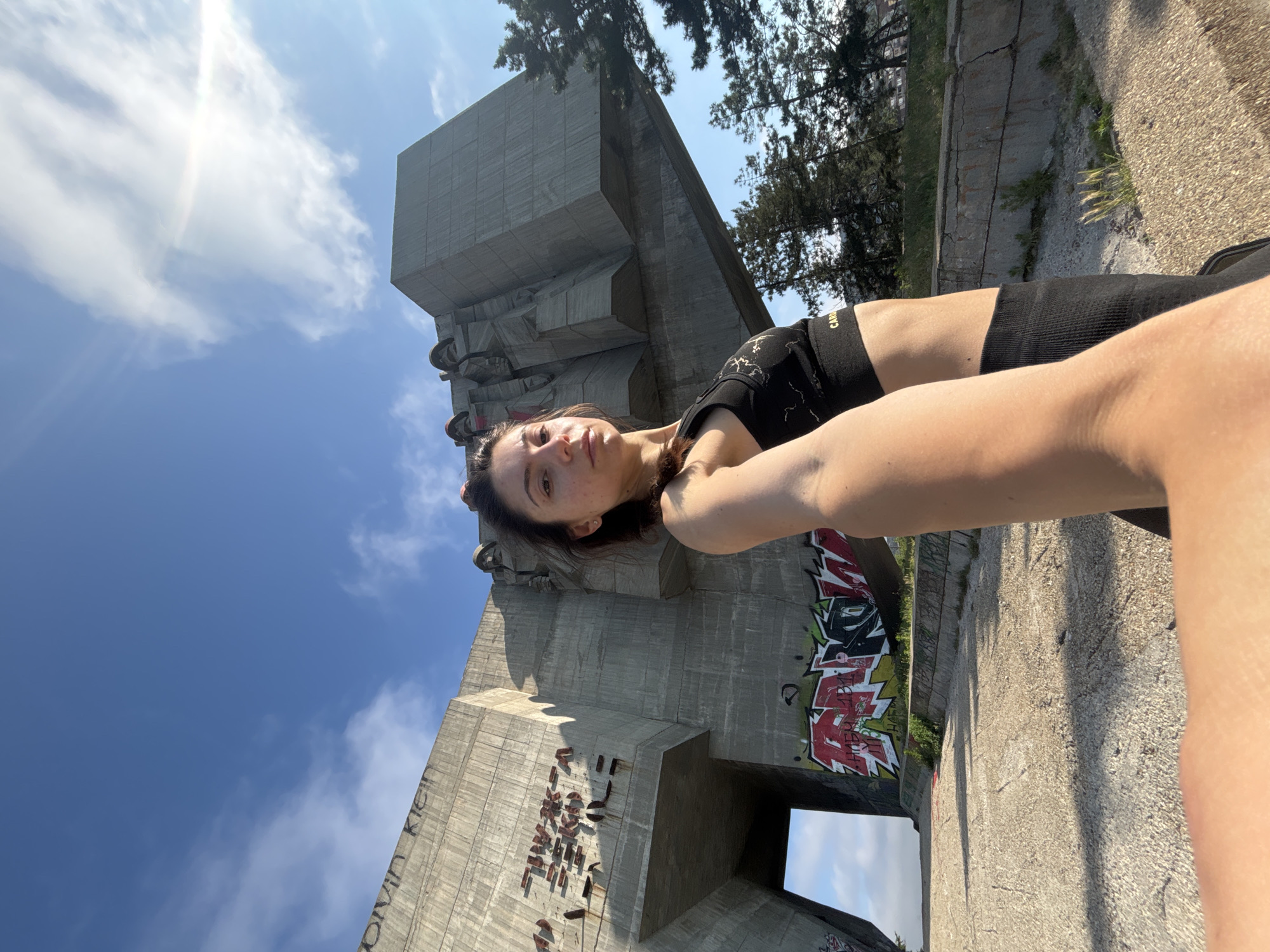 A woman is taking a selfie with a monumental concrete structure in the background. The woman is in the foreground, facing the camera and looking directly at it. She is wearing a black sports bra and black leggings. The concrete structure is large and imposing, with graffiti on its lower sections and some figures carved into its upper part. The setting appears to be outdoors, with a blue sky and some clouds visible. The overall impression is one of a modern art installation or a memorial site. This is the Buzludzha monument, a derelict monument in Bulgaria.