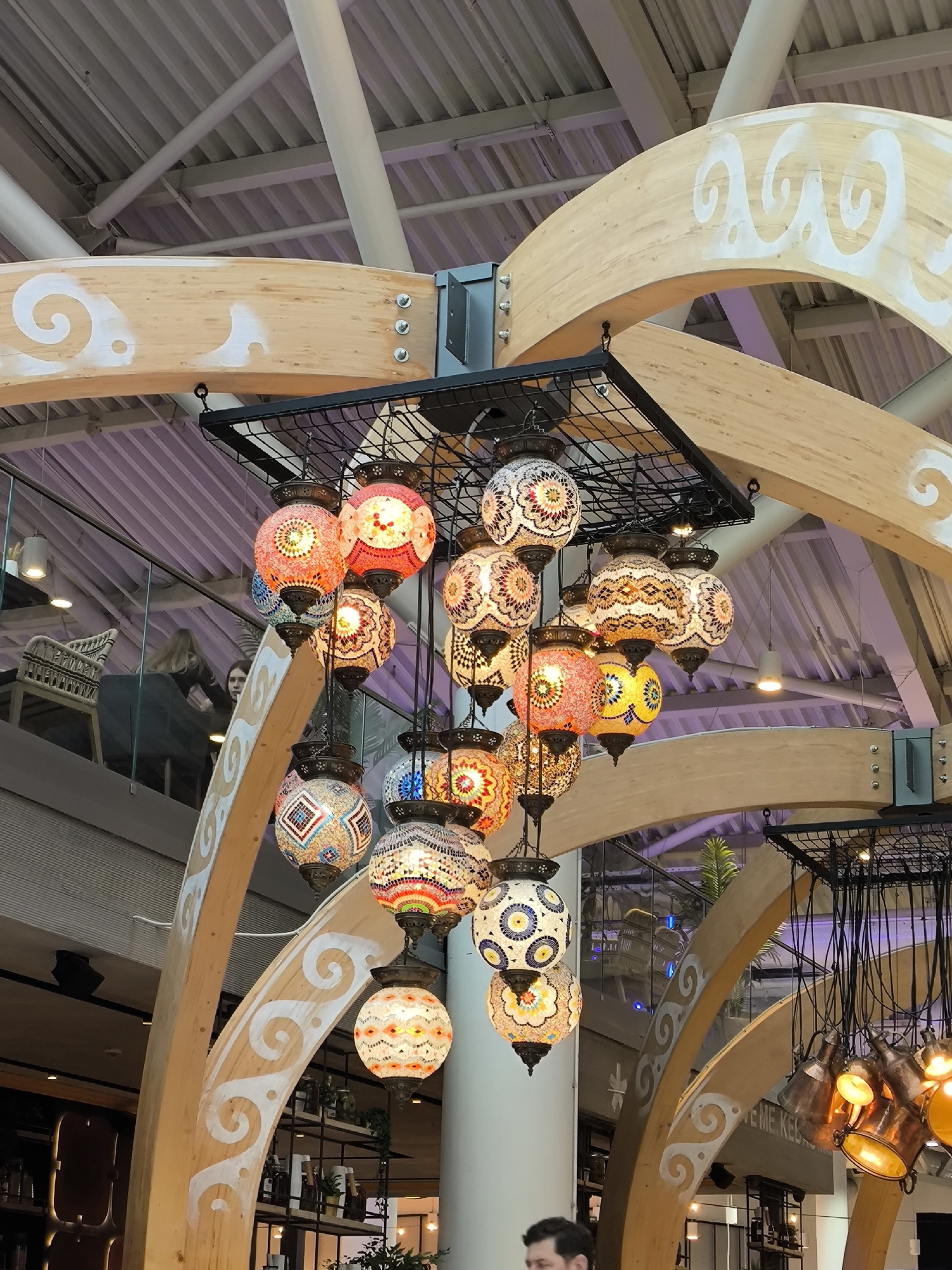 The image shows a decorative lighting fixture hanging from a black metal frame. The fixture is composed of multiple colorful, mosaic-style lanterns. The lanterns are suspended within a wooden, arched structure that is decorated with white, wave-like patterns. The structure is part of the interior design of a public space, likely a restaurant or shopping center. In the background, people can be seen on a mezzanine level, along with shelves and decorative items suggesting a commercial setting. The lighting and architectural details indicate an intentional aesthetic design. The city is unknown.