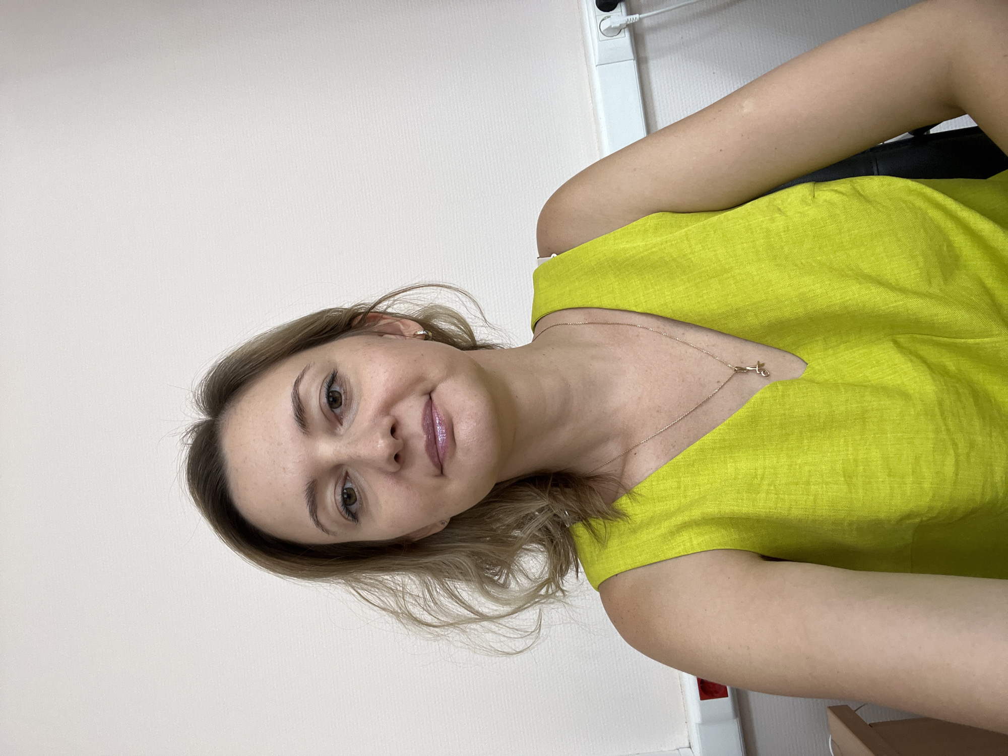 A fair-skinned woman with blonde hair and brown eyes is looking directly at the camera. She is wearing a lime green sleeveless V-neck top and a gold necklace with a small pendant. Her skin has some freckles. The background is a plain, light-colored wall with what appears to be a power strip with outlets on the right side. There is a hint of a desk on the lower left. The lighting suggests an indoor environment, possibly an office. The image was taken in Volgograd, Russia.