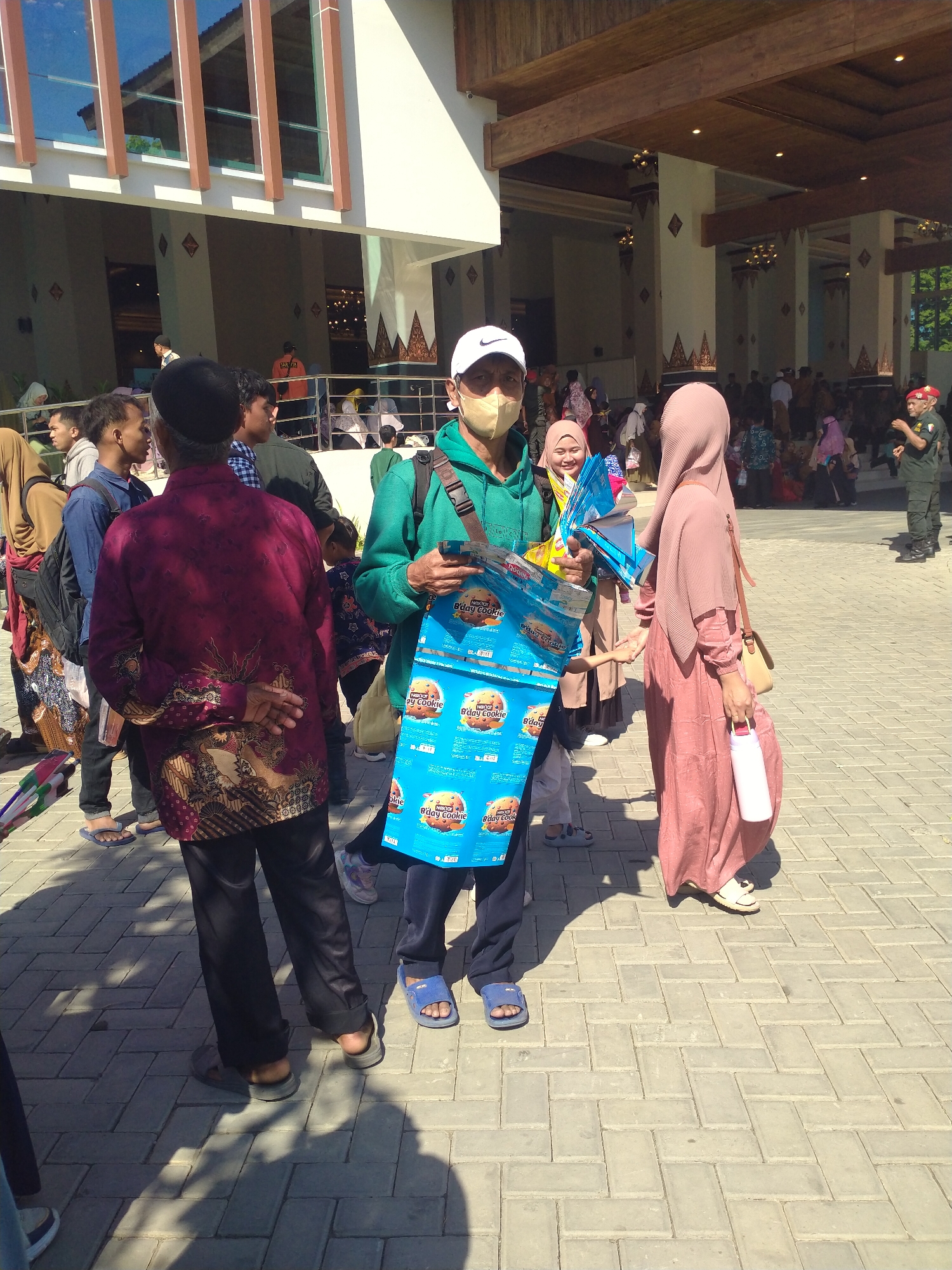This image captures an outdoor scene in Sukoharjo, Indonesia, likely in front of a building with a modern architectural style featuring large windows and wooden beams. In the foreground, several individuals are visible, some of whom appear to be engaged in social interaction.
A man in a green hoodie and white cap is holding a blue bag filled with what appear to be individually wrapped snack packages, possibly cookies, with text visible on the packaging that reads "B'day Cookie". He is wearing a light-colored mask. To his right, a woman in a pink hijab and a long, flowing pink dress is holding a white water bottle and has a light brown shoulder bag. She is smiling. A child in a light-colored hijab and a pink dress is standing near them, reaching towards the blue bag.
Further back, another man in a dark maroon batik shirt with intricate patterns is standing with his hands clasped behind his back. He is wearing black pants and flip-flops. Other people are seen in the background, some entering or exiting the building, suggesting a public gathering or event. The ground is paved with grey interlocking bricks. The overall atmosphere appears to be one of a casual daytime event.