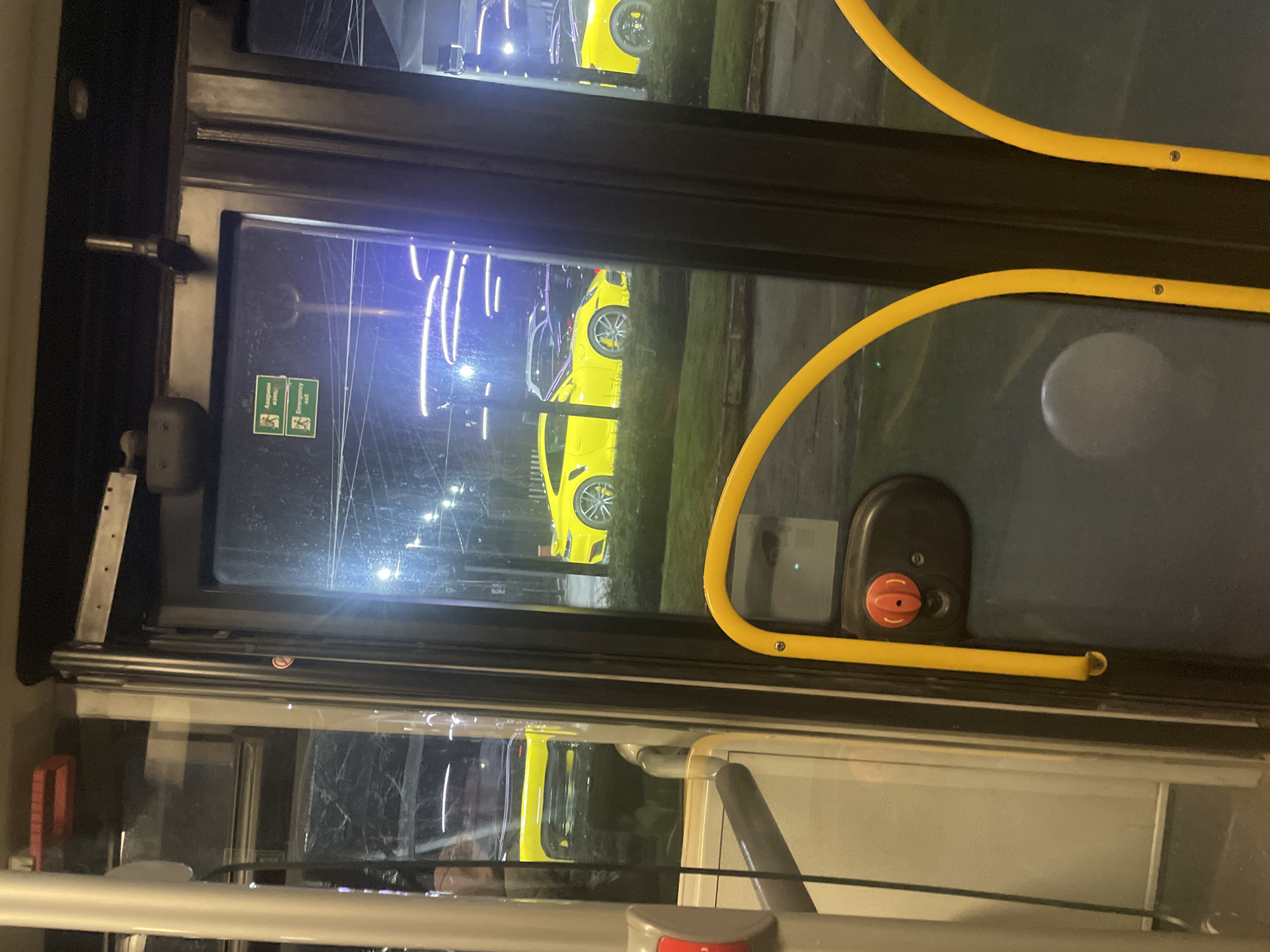 The image shows a view from inside a bus, looking out a window. Visible outside is a yellow Porsche parked on the street, with city lights in the background. The image was likely taken at night. The interior of the bus is visible, including a yellow handrail and an emergency exit sign on the window. The location is the city of Sofia, Bulgaria.
