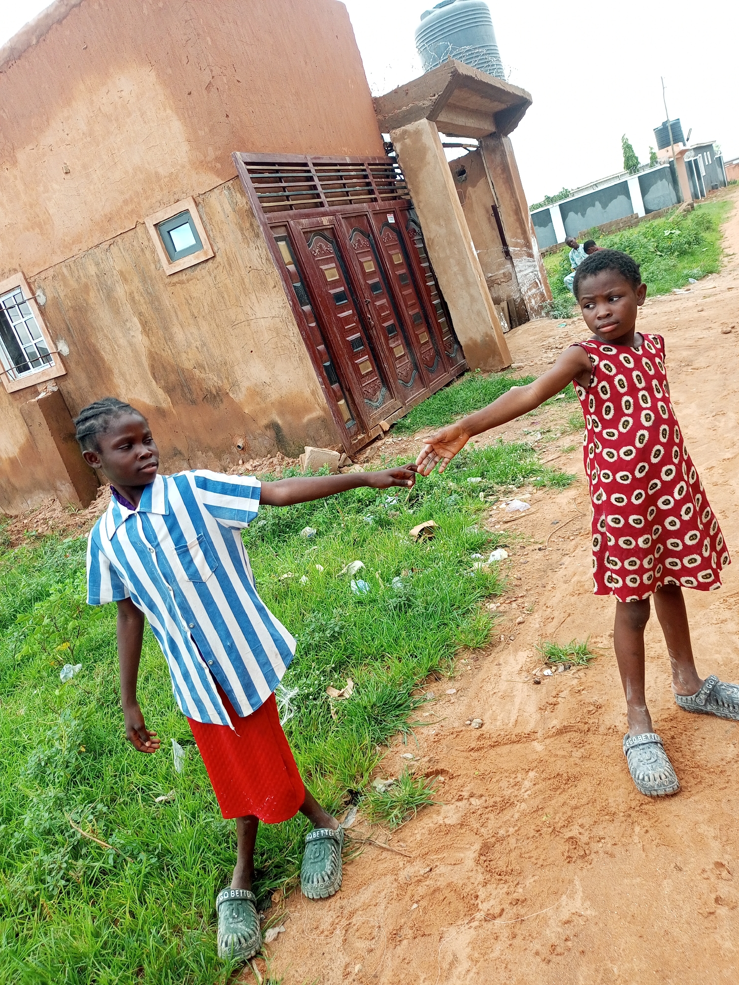 The image shows two young girls standing on a dirt path in front of a building with an orange, textured wall in Tempure, Nigeria. The girl on the left, wearing a blue and white striped shirt and a red skirt, extends her left hand towards the girl on the right. The girl on the right wears a sleeveless red dress with a white circular pattern and extends her right hand to meet the other girl's. Both girls are wearing sandals. The building behind them has a brown, wooden double door and a small, square window. Green grass grows to the left of the path. In the background, there are other buildings and a large water tank. The sky is overcast.