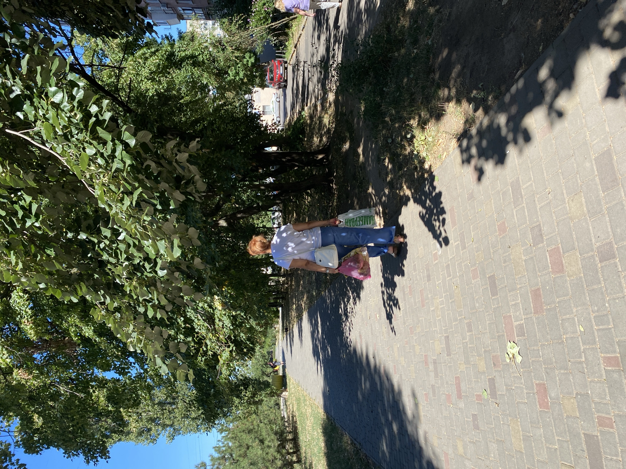 The image shows a sun-drenched pedestrian walkway in Chornomorsk, Ukraine, lined with trees. In the foreground, a woman with reddish-blonde hair walks away from the camera, carrying two plastic shopping bags. She is wearing a white t-shirt and blue jeans and a white crossbody bag. The walkway is paved with interlocking grey, beige, and reddish-brown bricks, with dappled sunlight and shadows from the overhead foliage creating a mosaic pattern. To the left, a grassy area is bordered by a line of conifer trees, and beyond that, a clear blue sky. To the right of the walkway, there is more grass and dense trees, with a paved road visible further in the distance where a red car and a silver car are parked. A multi-story building is visible in the background on the right. Further down the walkway, another person, possibly a child, is walking away from the camera, carrying a yellow object. The overall scene depicts a typical summer day in a public park or residential area.