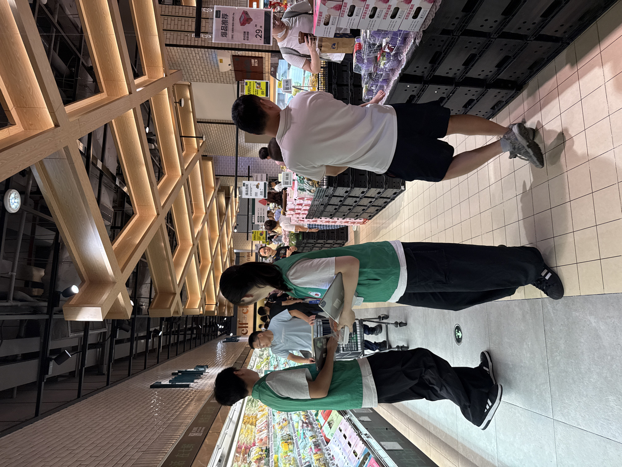 This media file shows a busy grocery store interior in Haizhu District, China. The scene is filled with people browsing and shopping. In the foreground, two young individuals wearing green vests and casual clothing are engaged in looking at products. One is holding a tablet. Behind them, other shoppers are seen pushing carts and examining merchandise. The store features modern wooden shelving and bright lighting. Visible text includes product pricing and promotional signs with Chinese characters, such as "爆品推荐" (popular item recommendation) and "龙泉驿水蜜桃" (Longquanyi peach). The overall atmosphere appears to be one of lively commerce. The time of day is not explicitly indicated, but the lighting suggests it is daytime or a well-lit interior. The weather is irrelevant as the scene is indoors. The emotions of the individuals are not clearly discernible, but the activity suggests a typical shopping experience.