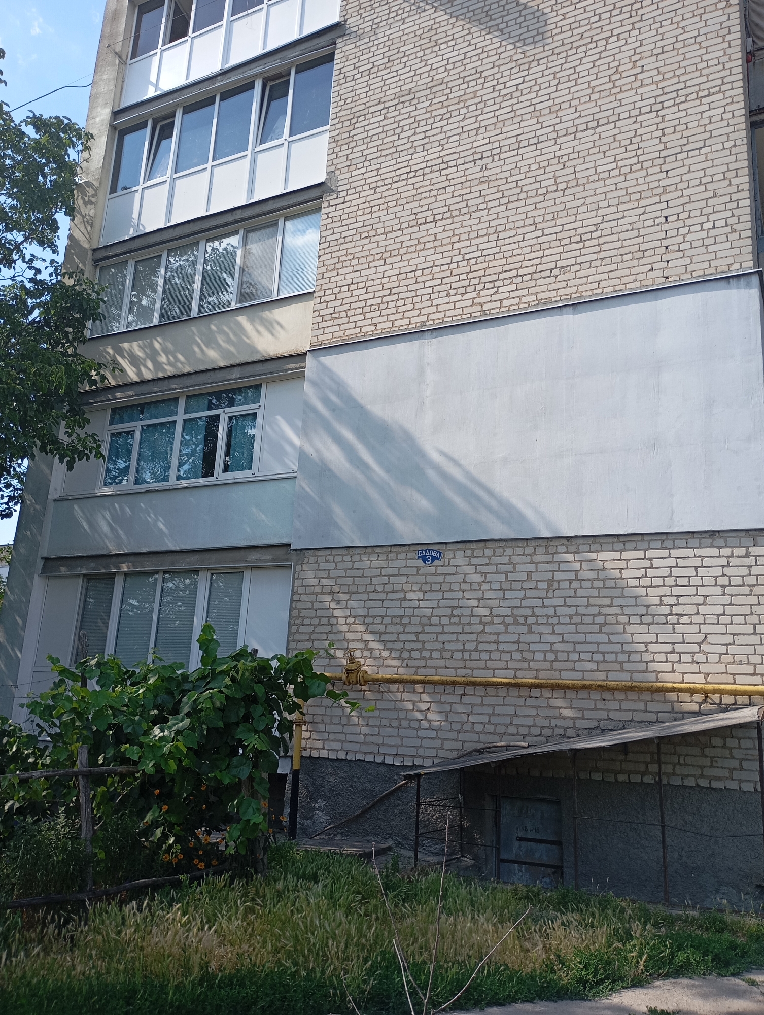 The image shows a portion of a multi-story apartment building in Sozonivka, Ukraine, during daylight. The building features a light-colored brick facade on its upper sections and lighter colored panels on the lower sections. Several windows and balconies are visible, some with white frames and glass. Green trees and foliage are present in the foreground and to the left of the building. A blue street sign with Cyrillic lettering and the number "3" is affixed to the lower brick section of the building. A yellow gas pipe runs horizontally across the lower brick section. Below this, there is a small, covered entrance with a metal door, surrounded by grass and a wooden fence. The sky is visible in the upper left corner, appearing clear and blue.