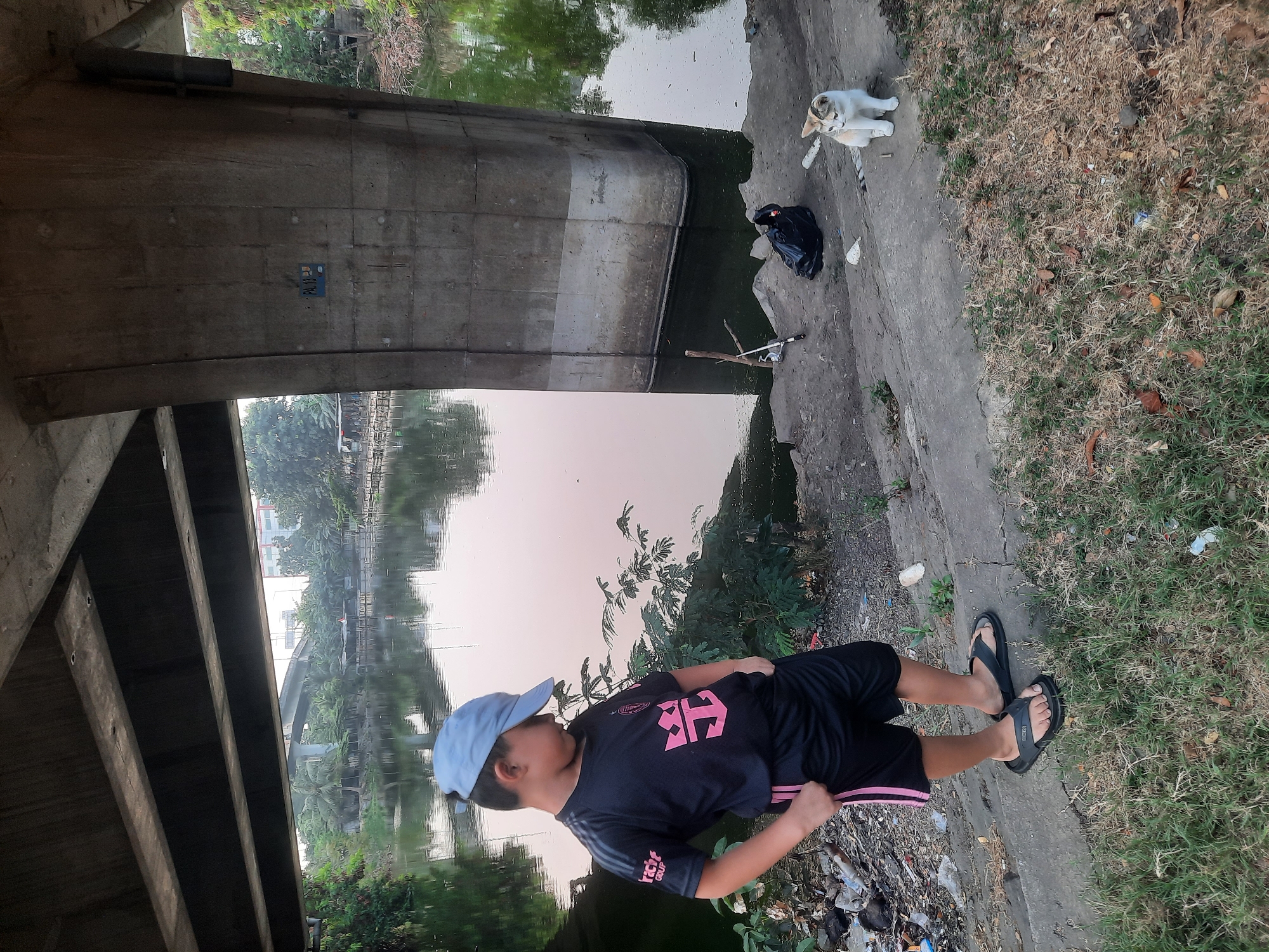 A young boy wearing a black t-shirt with pink trim and a blue baseball cap stands under a concrete overpass, looking down. He has his hands in his pockets. To his right, a small calico cat sits on the concrete ledge next to a black garbage bag and a fishing rod. The scene takes place near a canal or river, with a distant view of buildings and trees. The ground is covered in grass and debris. This location is under a flyover in North Jakarta, Indonesia.
