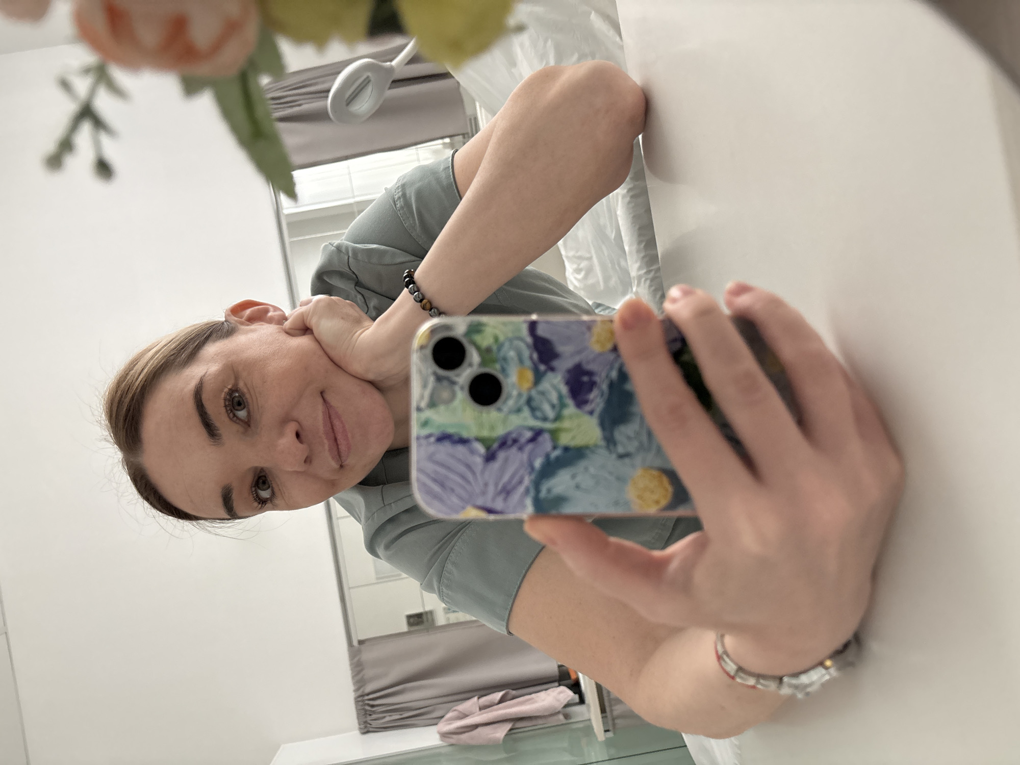 A woman is holding a phone and taking a selfie in a mirror. She is wearing a gray-blue uniform and has her head tilted to the side with her hand supporting her cheek. The phone has a floral-patterned case. A light and a flower are in the upper left corner of the mirror. Blinds are visible in the background. The setting appears to be a clinic or office. There is no definitive indication of the location being in the city of Romanovka, Russia.