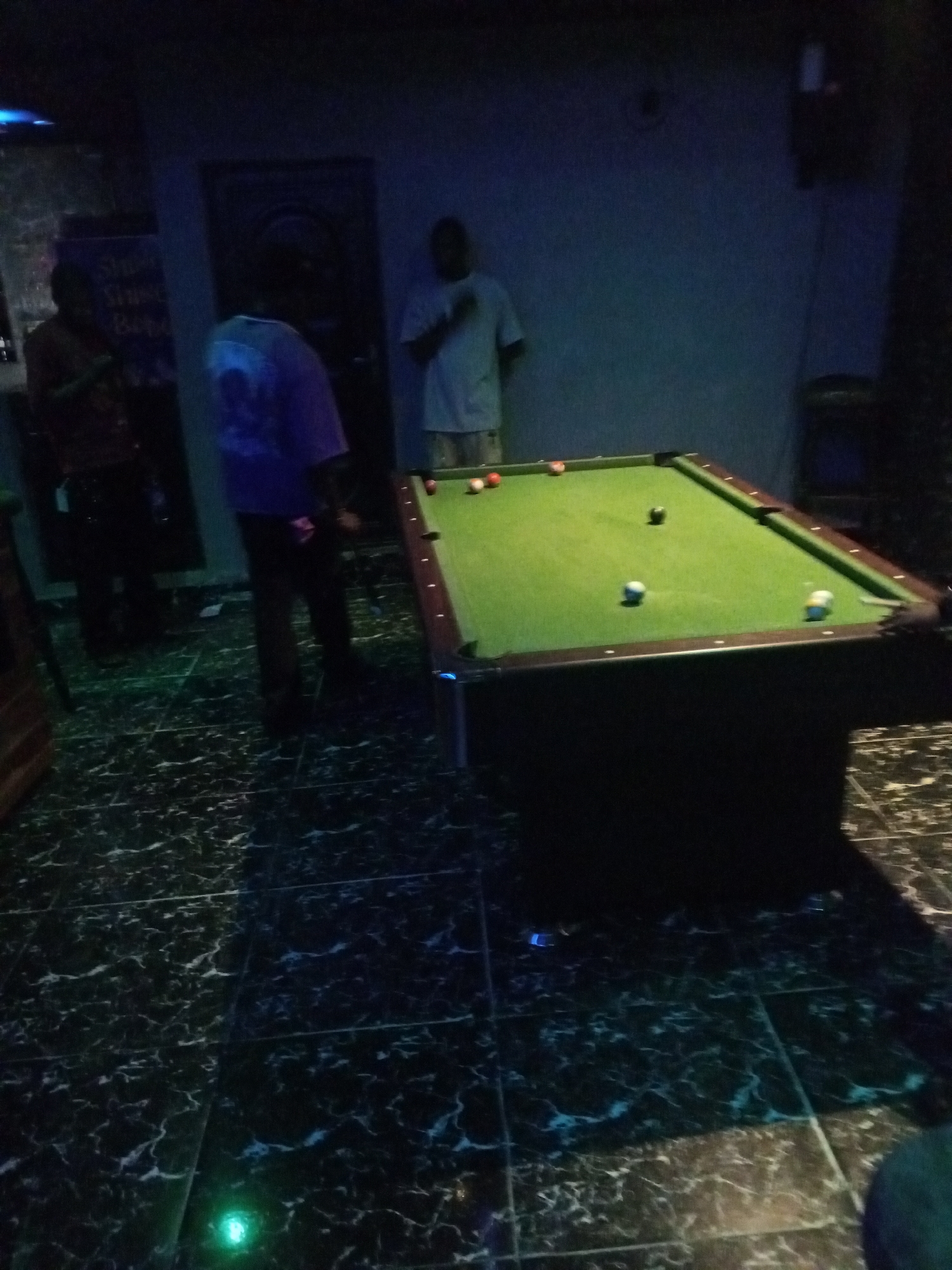 In an indoor environment with a dark, marble-patterned tiled floor and dark walls, three individuals are visible near a pool table. One person, wearing a light-colored t-shirt and shorts, stands at the far end of the pool table, facing towards the viewer. To their left, another person, wearing a blue t-shirt and dark pants, is positioned behind the pool table, looking towards the right. On the far left of the image, a third person, wearing a dark patterned shirt and dark pants, stands to the side, partially obscured by shadow, looking towards the pool table. Several pool balls are scattered on the green felt surface of the pool table. The lighting is dim, with blue and green lights visible in the scene, suggesting a bar or entertainment venue.