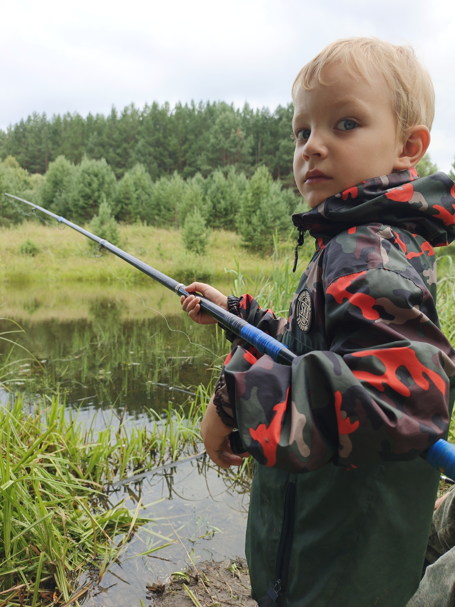 A young boy with blonde hair and blue eyes is fishing by a lake in Medvedyovka, Russia. He is wearing a camouflage jacket with red and green patterns. The boy is holding a fishing rod, and the line is in the water. Tall green trees line the lake in the background. The sky is overcast.