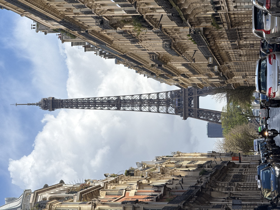 The image depicts a street scene in Paris, France, with the Eiffel Tower prominently centered in the background. The sky is partly cloudy with areas of visible blue. The street is lined on both sides by multi-story Haussmannian-style buildings constructed of light-colored stone, featuring ornate facades, balconies with black wrought-iron railings, and numerous windows. Some balconies display small green plants.
The paved street in the foreground and midground contains vehicular traffic. On the left, a line of parked or slowly moving cars includes several black taxis with illuminated "TAXI" signs on their roofs. On the right, a similar line of vehicles is present, featuring a silver Citroën C4 (license plate DL-716-AB) and a silver Volvo SUV. A single individual on a motorcycle, wearing a helmet and dark attire, is visible in the central lane. Bare-branched trees line the street in the midground, framing the approach to the Eiffel Tower. A modern, taller building is partially visible behind the base of the Eiffel Tower.