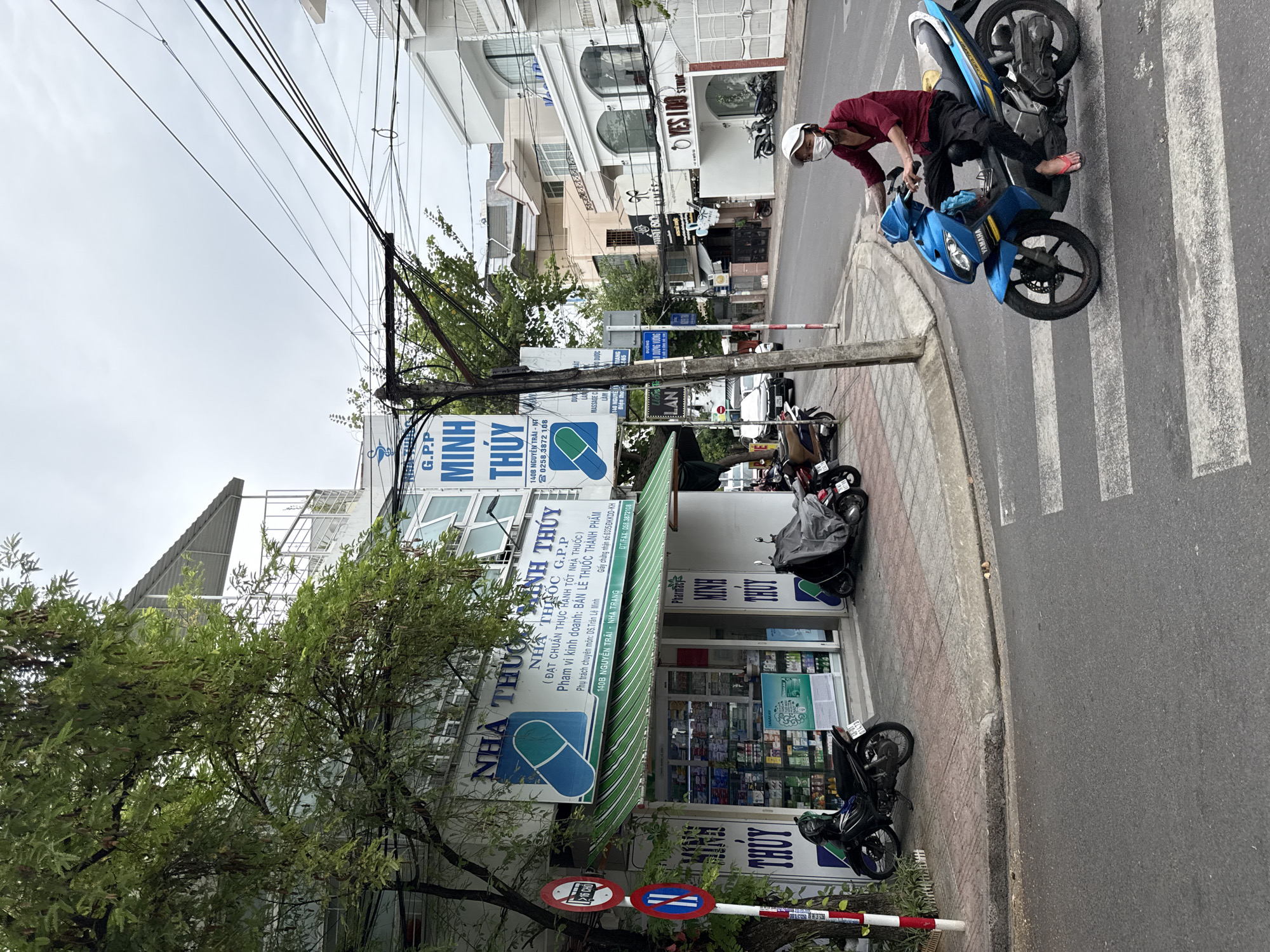 The image captures a street scene in Nha Trang, Vietnam. On the left, a pharmacy named "NHÀ THUỐC MINH THỦY" is visible, with its interior stocked with various pharmaceutical products. A blue and white awning extends over the shop entrance. Two motorcycles are parked on the sidewalk in front of the pharmacy. A traffic sign with red and blue circles and a diagonal line is posted on a pole near the pharmacy. In the center of the frame, a person wearing a white helmet and a face mask is riding a blue Yamaha motorcycle across a pedestrian crossing. The person is wearing a red shirt and black pants. The street is paved, and buildings line the opposite side of the road, featuring a mix of architectural styles. Overhead, a complex network of electrical wires is suspended on poles. The sky is overcast.