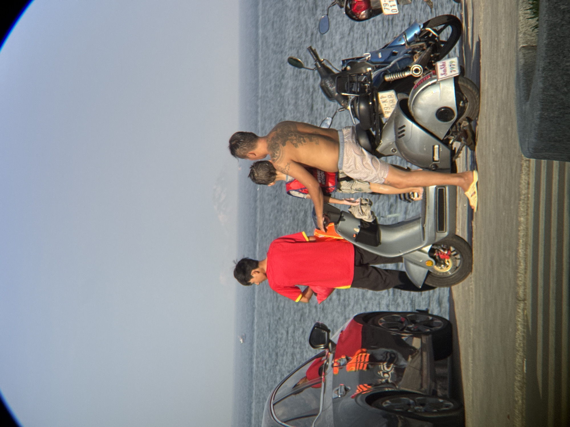 In Nha Trang, Vietnam, a man with a prominent back tattoo is seated on a silver scooter, facing away from the camera. He is wearing light-colored shorts and yellow flip-flops. Beside him, a young boy wearing a red life vest stands next to the scooter. To the left, a person wearing a red shirt and black pants stands near a dark-colored car. The car's reflection shows a person wearing an orange life vest. The scene is set on a paved area next to a body of water, likely the sea, with a hazy sky and distant landmass visible. Another scooter, predominantly blue, is parked behind the silver scooter, and a third scooter's rear is visible to the right. The overall atmosphere suggests a casual outing near the coast.