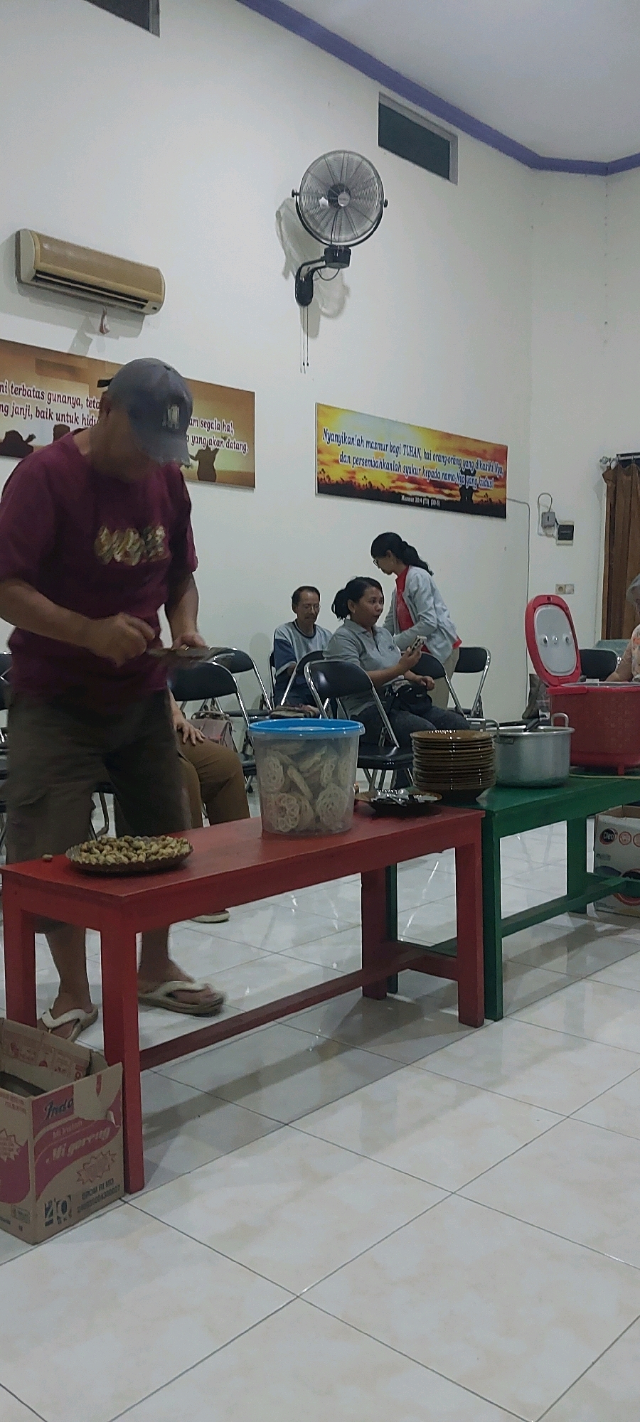 The image shows a gathering of people indoors, likely in a community hall or similar venue in Keling, Indonesia. In the foreground, a man wearing a maroon t-shirt and a baseball cap is standing in front of a red table. He is holding something in his hands and appears to be interacting with it. On the red table, there is a plate of peanuts and a clear plastic container filled with crackers. Behind the man, several other people are seated on chairs, some of whom are looking at the man or at their phones. A woman in a gray shirt is seated and holding a mobile phone. Another woman in a light-colored jacket is standing and interacting with the seated woman. The room has white walls with posters displayed on them. One poster has text in Indonesian and a sunset illustration. Another poster shows text in Indonesian with a silhouette of people looking at a sunset. A wall-mounted fan is visible on the wall, along with an air conditioning unit. Further back, there are green and red tables with stacks of plates and a pot. The floor is tiled. The overall scene suggests a casual social gathering or event.
