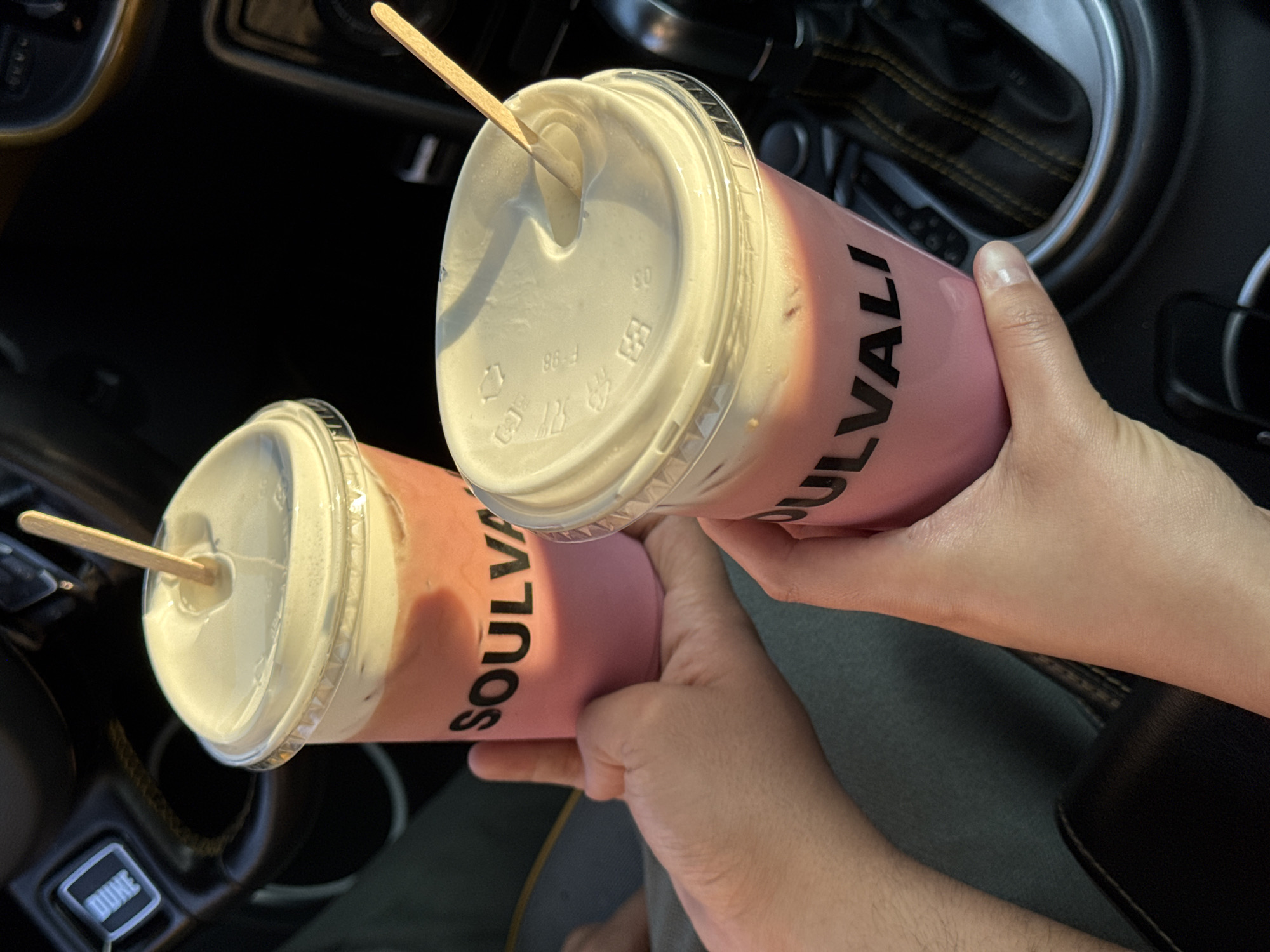 Two pink Soulvali brand drinks with white lids and wooden stir sticks are being held in someone's hands inside a vehicle. The vehicle's dashboard and gear shift are visible. The location is likely Puebla, Mexico, given the brand name on the cups.