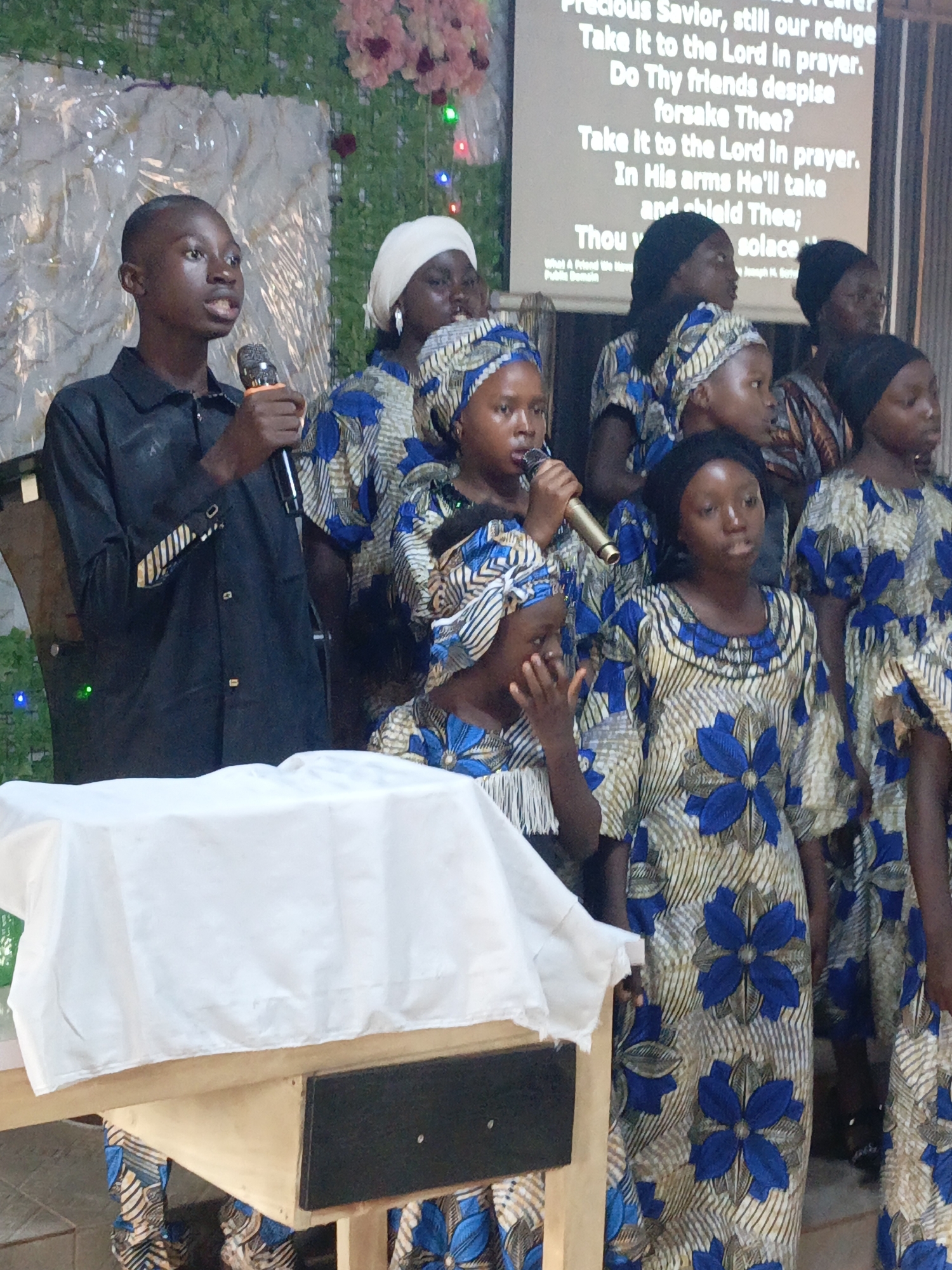A group of people, including children, are standing on a stage in Akwanga, Nigeria. The group is dressed in matching outfits patterned with blue and tan flowers. One young man on the left holds a microphone and appears to be speaking or singing. Another person is speaking into a microphone in the center of the group. Behind them is a screen displaying text from a hymn. The stage is covered with a white cloth-draped table in front. Behind the screen is a floral decoration.