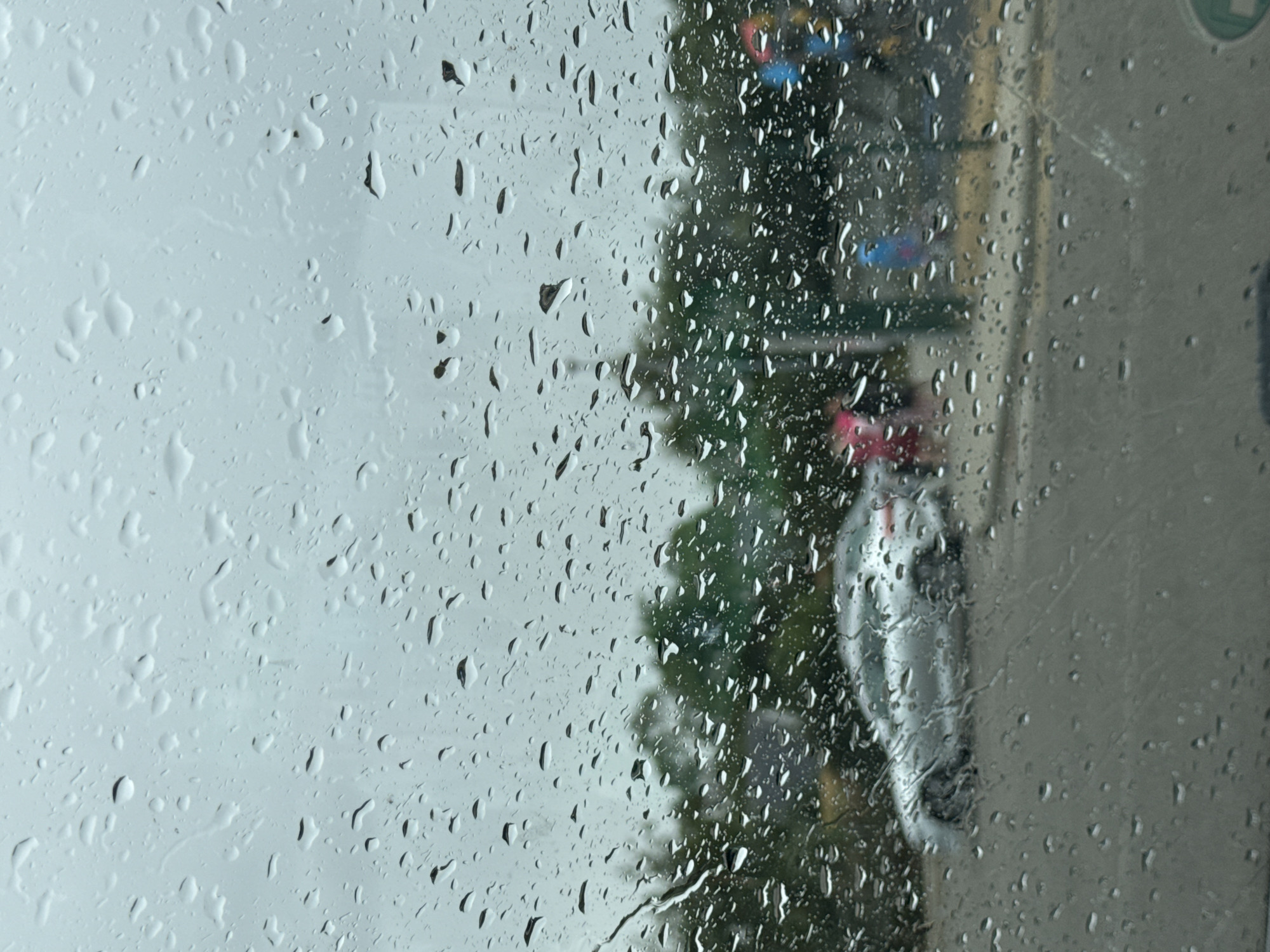 The image depicts a view through a rain-streaked car window, looking out onto a street scene in Courtown, Ireland. The sky is overcast and the weather appears to be rainy. In the mid-ground, a silver car is visible, parked or driving slowly on the asphalt road. Behind the car, a line of trees forms the backdrop. Further in the background, the indistinct shapes of buildings and possibly a traffic light or signal can be seen, with flashes of color, one appearing red and another blue. The foreground is dominated by numerous raindrops clinging to the car's window, distorting the view of the outside. The overall impression is one of a damp and gray day in a suburban or town setting in Courtown.