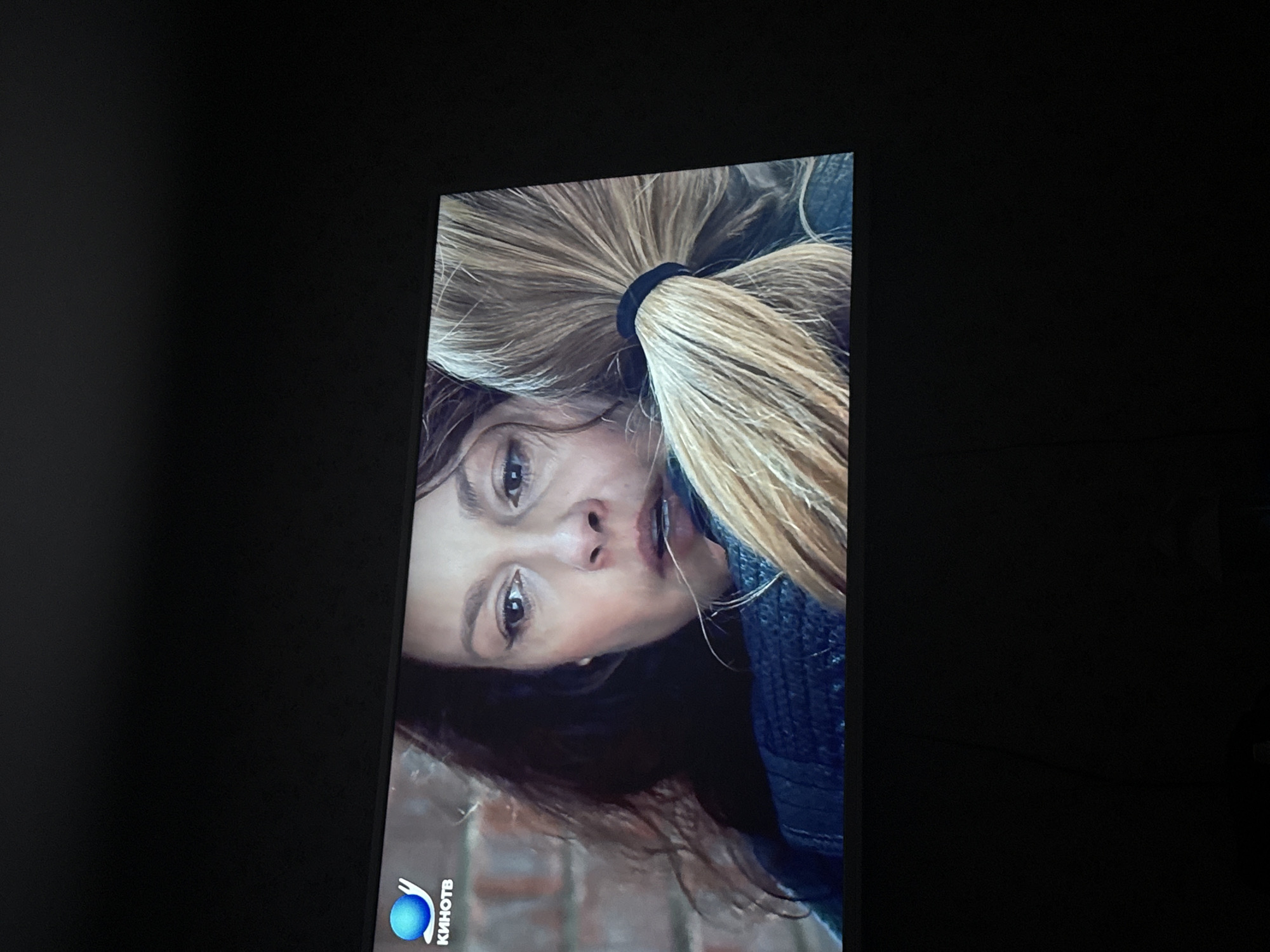 The image shows a television screen displaying a close-up of a woman's face. She has fair skin, light brown hair pulled back into a ponytail with a black elastic band, and wears a dark jacket. Her facial expression is slightly downturned. The screen also displays the logo "КИНОТВ" (KINOTV), a Russian-language television channel. This suggests the image is likely from a TV show or movie aired on that channel. The black background indicates the television is surrounded by darkness. There is no way to determine the location of Mytishchi, Russia, based on the image alone.