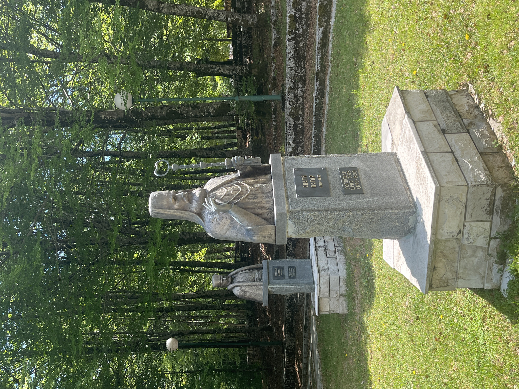 This outdoor scene depicts two statues situated near a grassy area and a backdrop of trees in Velji Bostur, Montenegro. The statues, mounted on stone pedestals, are likely commemorative monuments. The foreground includes green grass and the base of the statue's pedestal, constructed with light-colored stones. The statue depicts a figure holding what appears to be a staff with an ornate end. Further back, another statue is visible, though smaller and partially obscured. Dense trees fill the background. The lighting suggests a sunny day, casting shadows across the grass and statues.