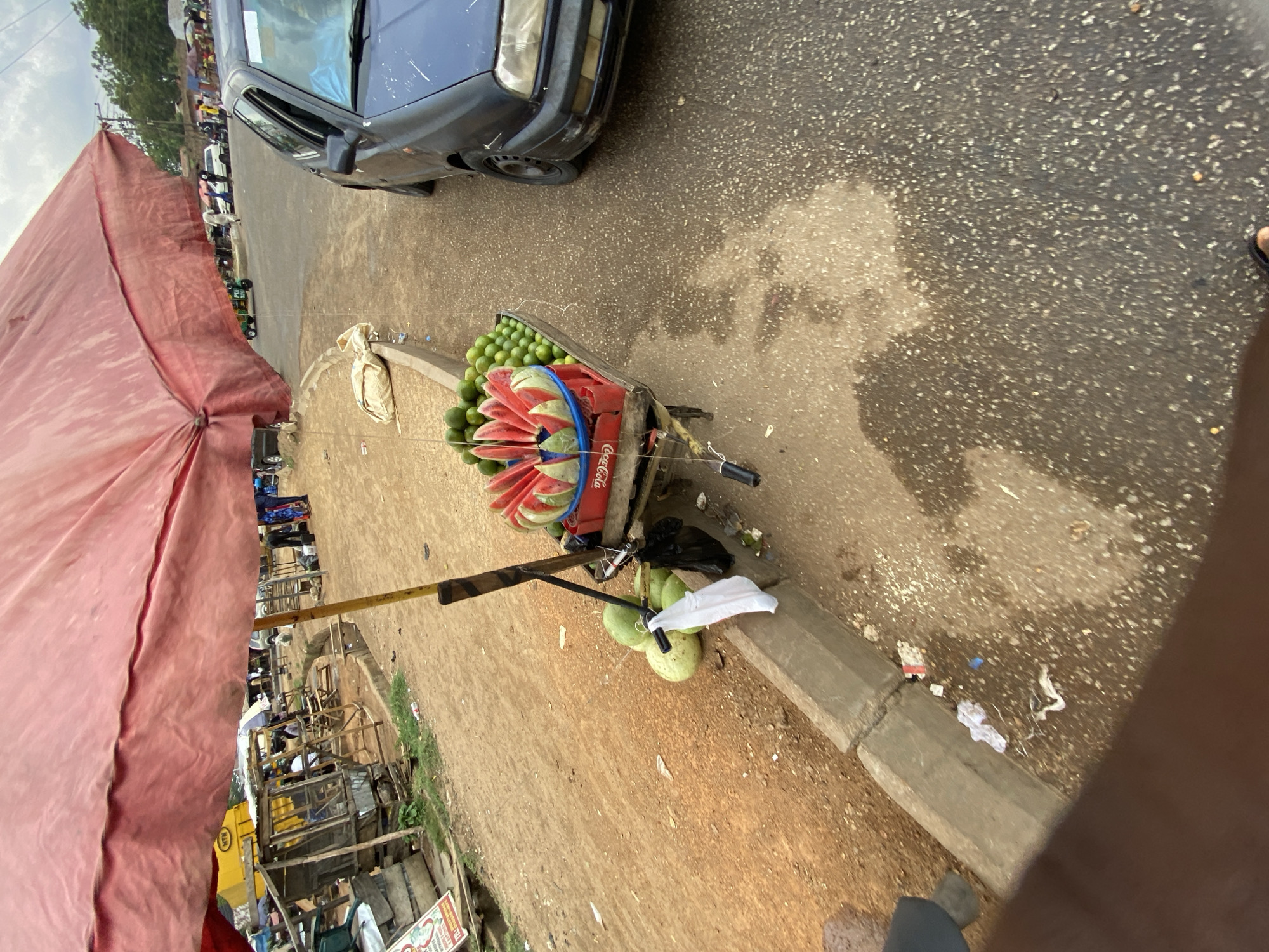 The image shows a street scene in Bukuru, Nigeria. A blue car is parked at the roadside. A vendor's cart, heavily laden with watermelons and green citrus fruits, is parked on the curb. The cart is labeled with "Coca-Cola." A large, red umbrella provides shade for the vendor's stand, which is set up amidst other market stalls. In the background, other vendors and pedestrians are visible. The road is paved and partially wet. There is trash visible on the curb.