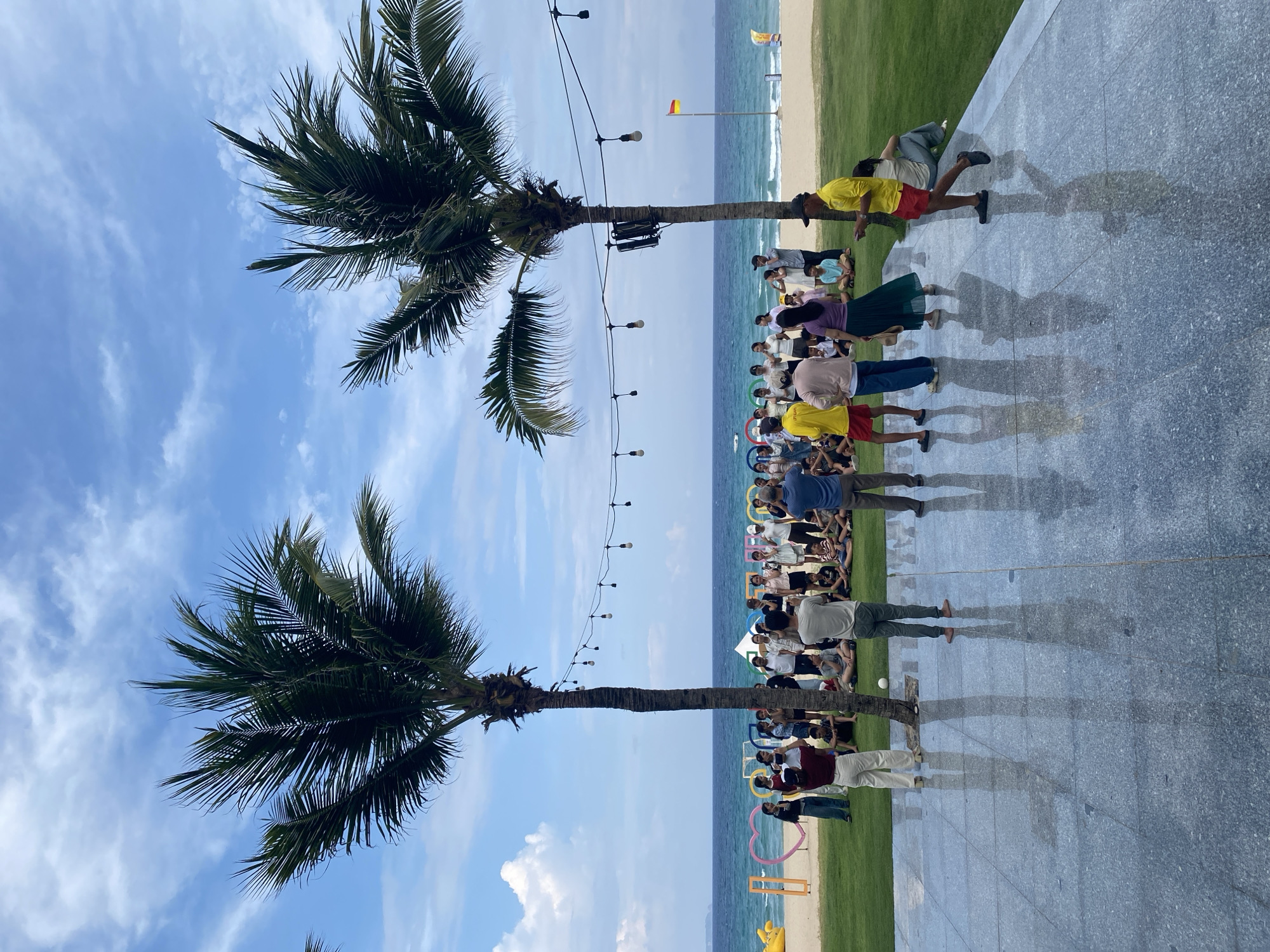 The scene depicts a group of people gathered outdoors at a beachfront location in Bắc Cam Ranh Ward, Vietnam. Two palm trees frame the image under a partially cloudy blue sky. A string of lights spans between the trees above the group. A lawn stretches toward the beach and ocean in the background, where a flag is visible. The group seems to be posing or watching something. Several individuals are visible along a paved surface that extends into the foreground, casting reflections of the people and the surrounding environment. A large, colorful sign with shapes and letters stands on the grass.