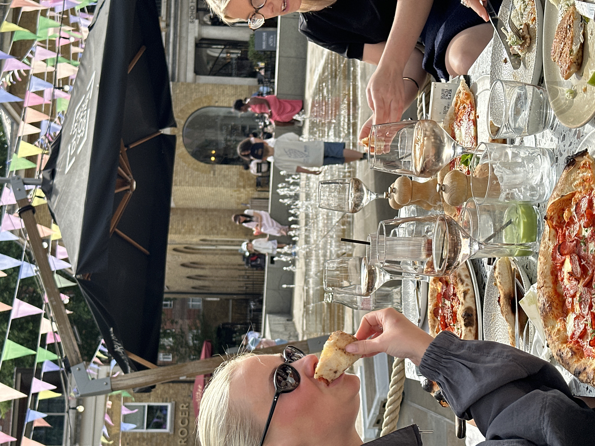 The image depicts a scene at an outdoor restaurant in Soho, London. A blonde woman wearing sunglasses is holding a piece of pizza to her mouth. On the table, there are pizzas, glasses filled with liquid, salt and pepper shakers, plates, and silverware. Another person is seated across from the woman, wearing glasses and a black top, and is eating. In the background, a fountain is visible with people walking by. Colorful triangle flags are strung overhead and a black umbrella provides shade. The architecture in the background includes a building with the word "SOHO" visible at the top.