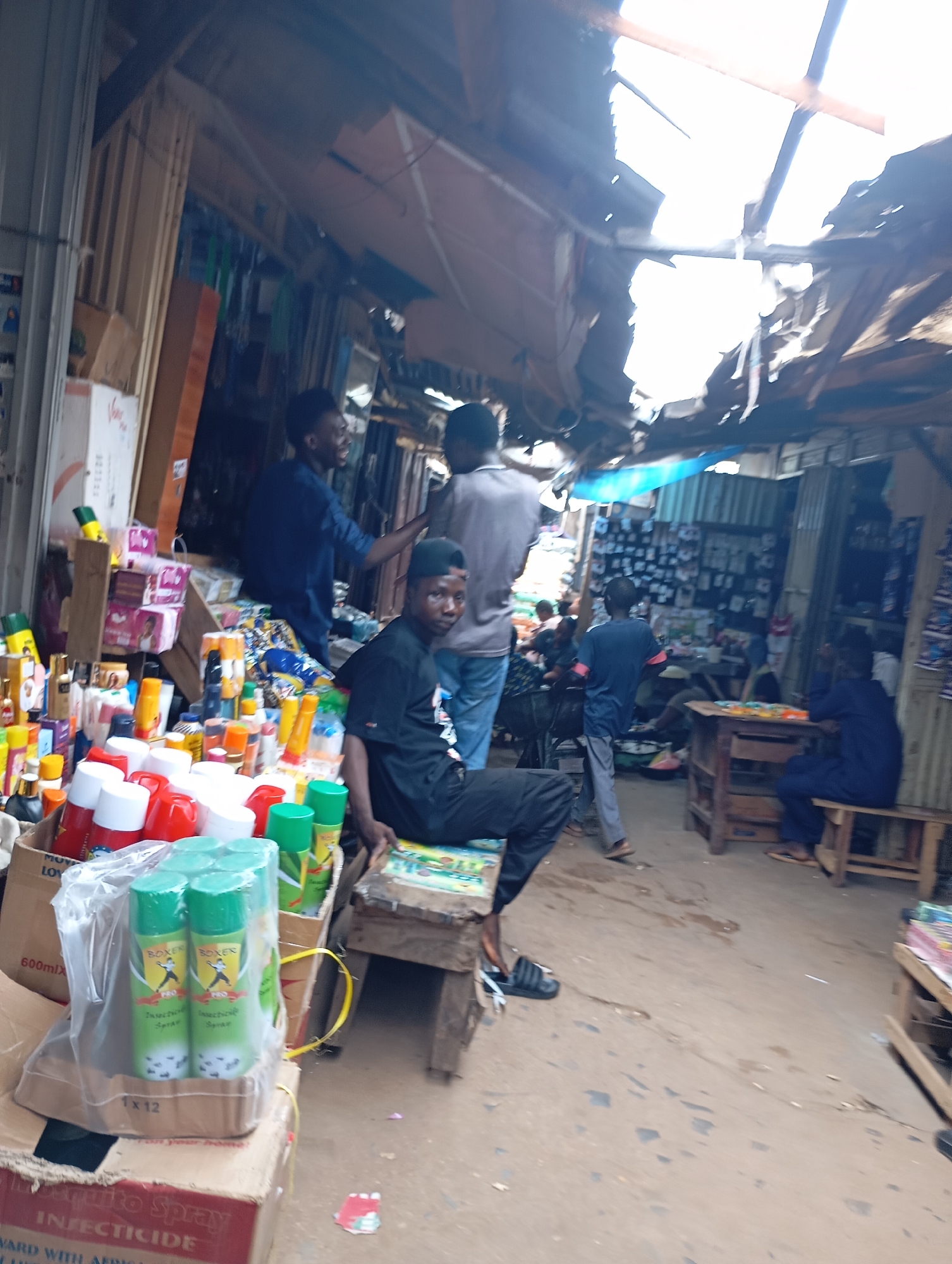 This image depicts a bustling outdoor market scene in Shendam, Nigeria. A group of individuals are engaged in commercial activities, with some standing and others seated. Several market stalls are visible, displaying a variety of products including what appear to be insecticides, toiletries, and other packaged goods. The stalls are constructed from various materials, with makeshift roofing structures, suggesting a temporary or semi-permanent market setup. The overall environment appears to be a typical marketplace in a Nigerian city, with an emphasis on small-scale trade and a lively atmosphere. The image captures the essence of everyday commerce in Shendam.