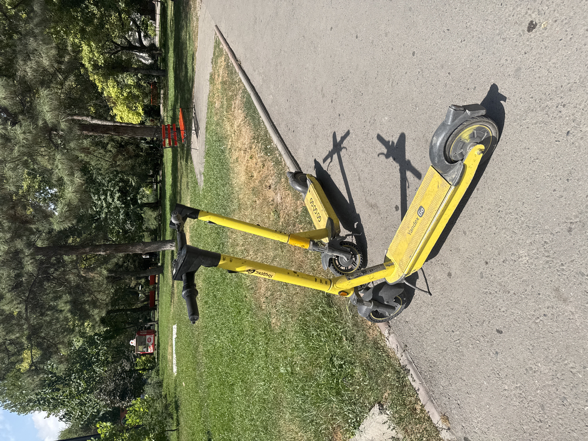 Two yellow electric scooters, identified by "Yandex Go" branding, are parked on an asphalt path in a park setting. The scooter in the foreground is positioned closer to the viewer and is angled slightly to the left, while the second scooter is behind it and to the right. Both scooters are upright, suggesting they are parked and not in use.
The park features lush green grass on the left side of the image, with a paved walkway running through it. Beyond the walkway, there are numerous trees, casting dappled shadows on the ground. In the background, a red park bench is visible under the shade of trees, and further back, a small red structure that appears to be a kiosk or cart can be seen. The overall environment suggests a sunny day in a recreational urban area. The scene is devoid of any visible people.