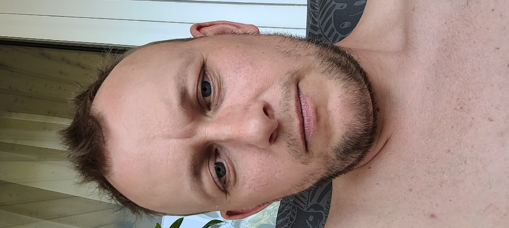 A close-up, forward-facing selfie of a Caucasian man with thinning brown hair, blue eyes, and stubble. He is looking directly at the camera with a neutral expression. The man is outdoors, with a blurred background of vertical blinds and some greenery visible to the left. The image appears to have been taken on a patio or balcony, possibly in a residential area of Heusenstamm, Germany, given the architectural style of the blinds and the overall setting. The man is shirtless, and the upper part of his chest is visible. The lighting suggests it is daytime.