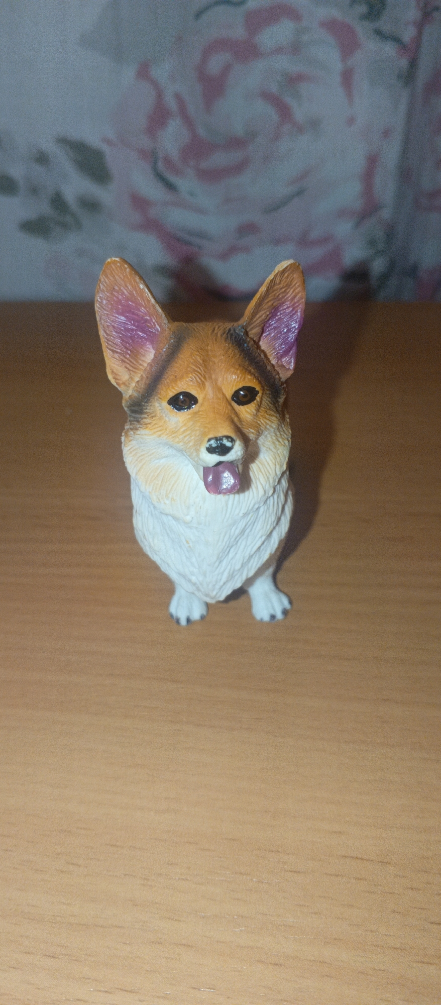 A toy corgi dog stands on a wooden table. The toy is brown and white with purple accents inside its ears. Its tongue is sticking out. The background is a blurry pattern of pink and grey floral shapes. The lighting is dim, casting a shadow behind the toy. The scene is indoors and the location is not specified beyond "Unknown, Unknown".