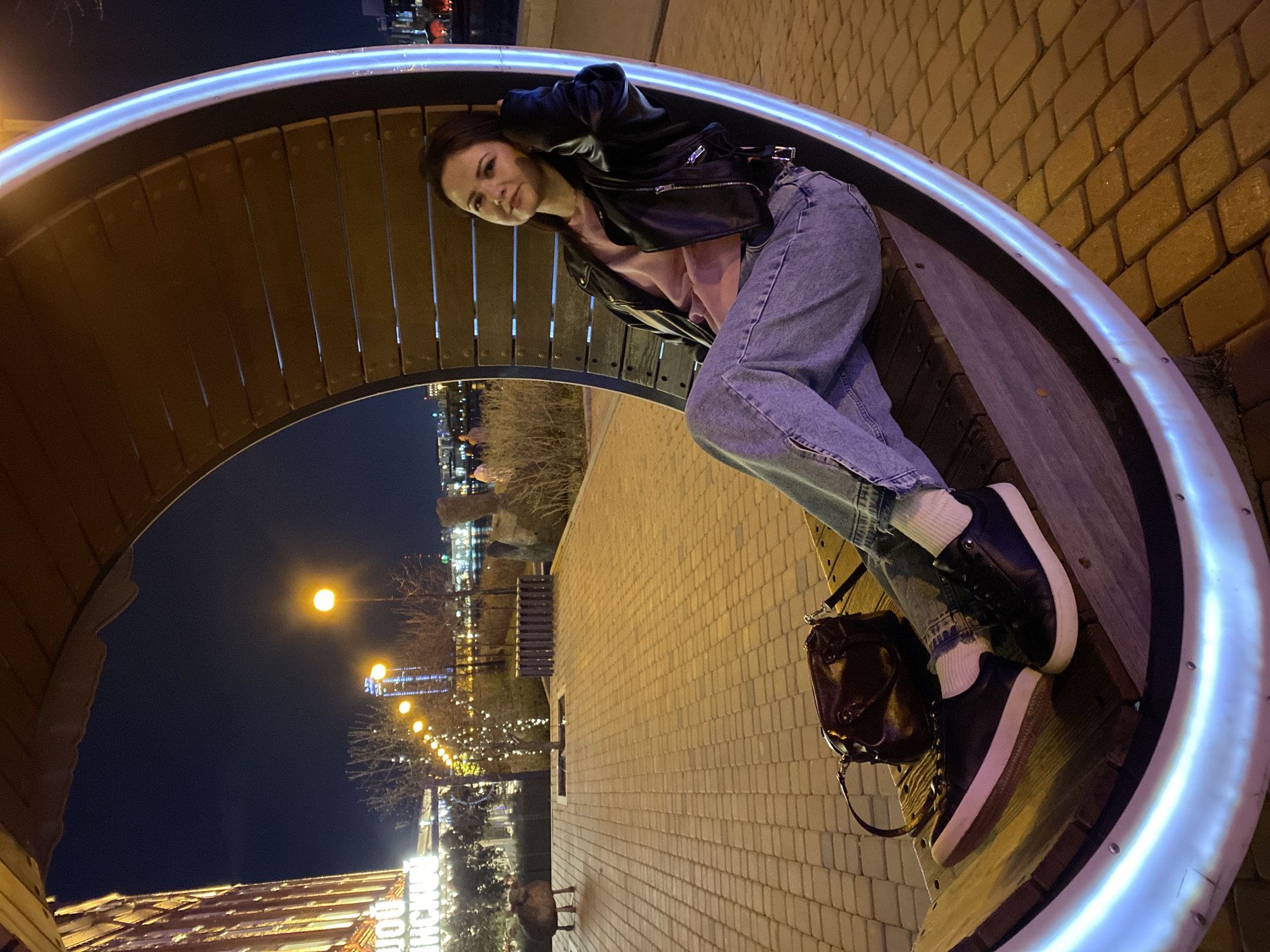 A woman wearing a black leather jacket and blue jeans sits on a curved bench with integrated lighting. She also wears a pink shirt and black sneakers with white soles. Her bag sits next to her. The bench is located in Yekaterinburg, Russia, with city lights visible in the background at night. The ground is brick.