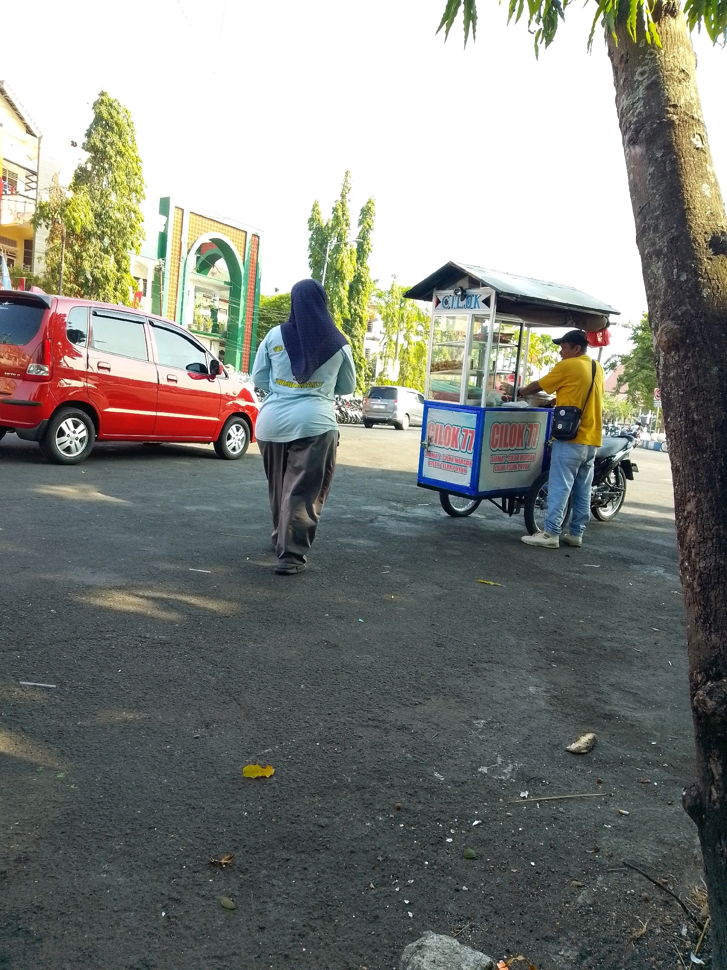 A wide shot captures an outdoor scene in Jember, Indonesia, with a clear sky. In the foreground, a woman wearing a dark hijab and a light blue long-sleeved shirt with dark gray pants walks away from the camera, her back towards the viewer. To her right, a man in a yellow t-shirt, dark cap, and blue jeans stands behind a mobile food cart. The cart, primarily white and blue, is adorned with text and graphics. The man appears to be serving or preparing food. A red car is parked to the left of the frame. In the background, a distinctive arched gateway structure with green and beige accents stands prominently. Trees with lush green foliage are visible throughout the scene, and a large tree trunk is on the right side of the image. The ground is paved.