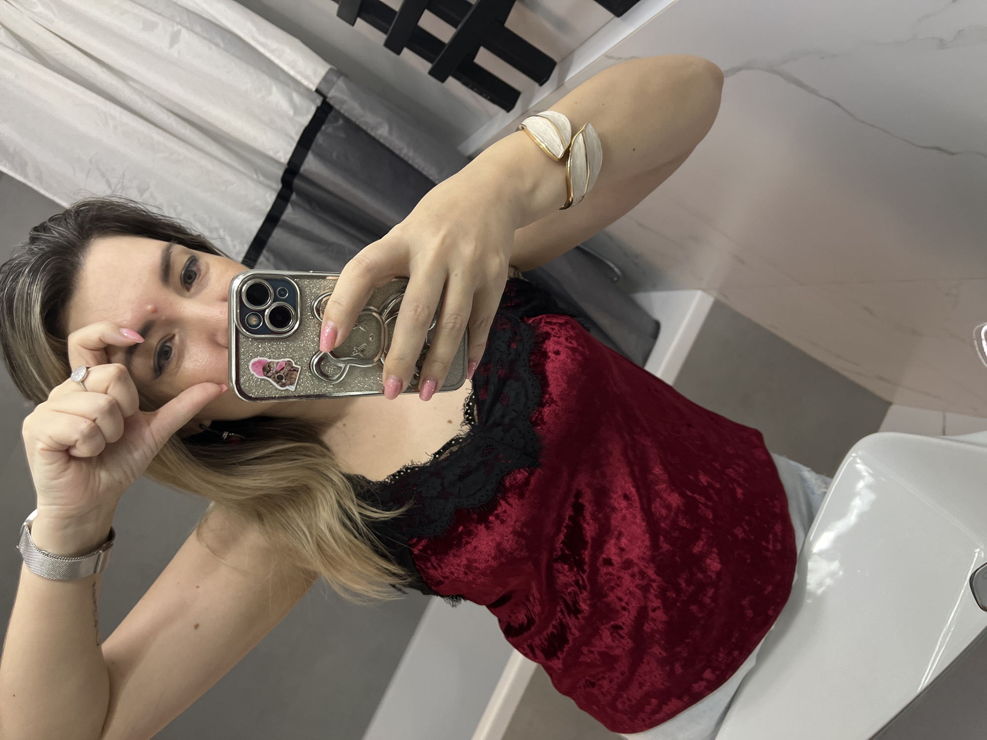 A young woman is taking a selfie in a bathroom. She is holding up a phone with a sparkly case, and her face is partially obscured by her hand. She is wearing a red velvet camisole with black lace trim. On her left wrist, she has a silver watch and a gold and white bracelet. Her nails are painted pink. The background shows a shower curtain and a marble countertop. The setting appears to be a modern bathroom. The image is well-lit, suggesting it was taken during the daytime. The woman's expression is playful as she looks into the phone's camera.