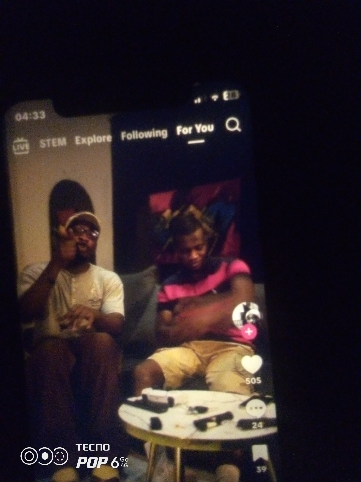 The image shows a smartphone displaying a TikTok-like interface with two men seated. The man on the left is wearing a beige t-shirt and a cap, and he is gesturing with his hand. The man on the right is wearing a pink and black striped shirt and yellow shorts. A small round table with objects on it is visible in the foreground. The scene appears to be indoors, possibly a living room, in a dimly lit environment. The time stamp on the phone indicates "04:33." The phone also displays "LIVE," "STEM," "Explore," "Following," and "For You," along with icons for likes, comments, and shares. The bottom of the phone displays "TECNO POP 6 Go 4G". There is no indication of the city of Howayi, Nigeria within the image.