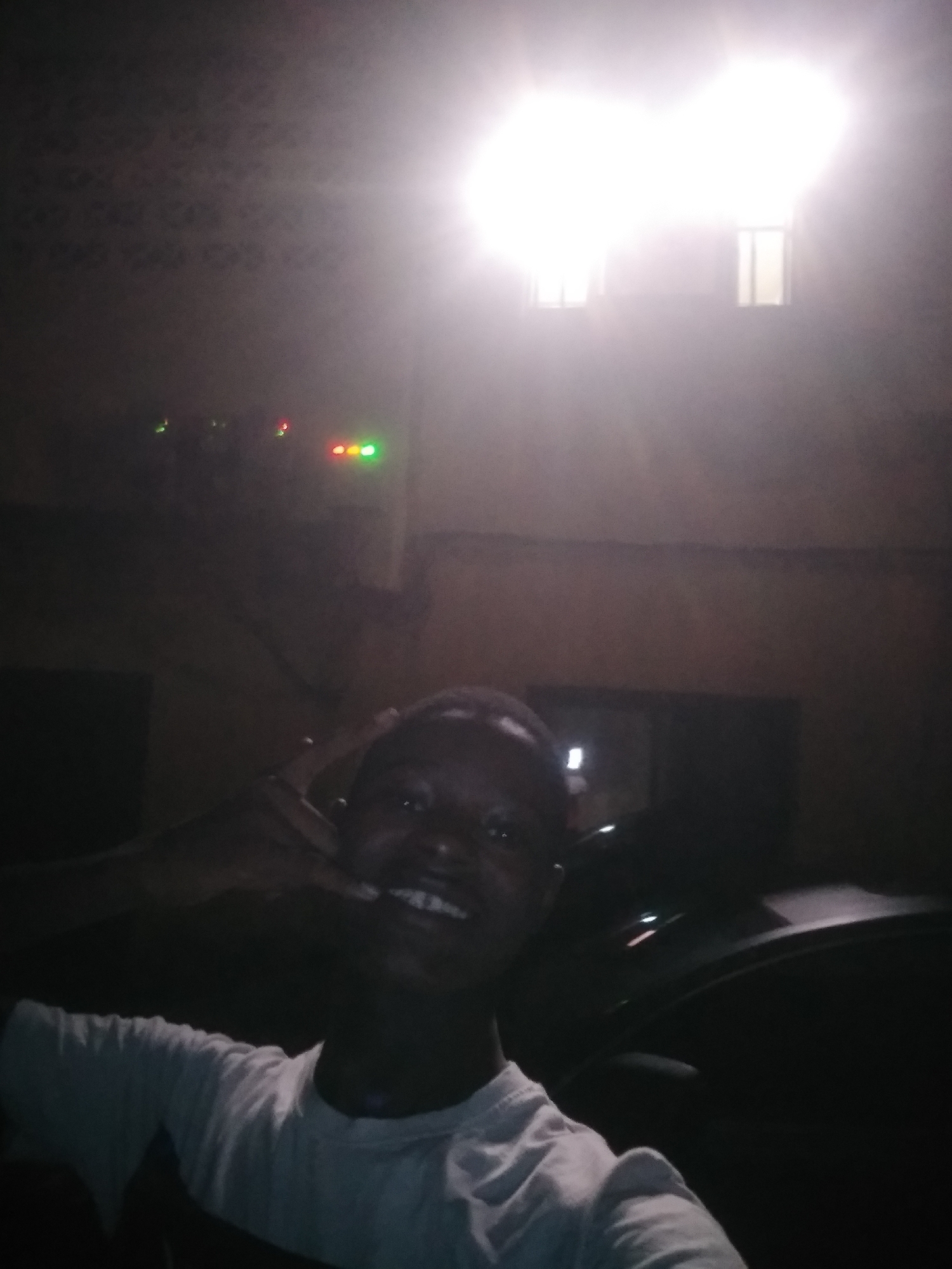 The image captures a young person smiling in a dimly lit environment at night in Shangisha, Nigeria. The individual, positioned centrally in the foreground, is wearing a light-colored (likely white) t-shirt and appears to be in a cheerful mood, grinning broadly with visible teeth. Their right hand is raised to their mouth, possibly holding something or making a gesture.
The scene is dominated by an extremely bright, overexposed light source in the upper center, creating a significant lens flare and obscuring details. Behind this intense light, two faint rectangular shapes resembling windows are barely visible. To the left and below the main light source, several small, colored indicator lights are visible: some green and red lights clustered together, and further right, a set of three lights (yellow, red, and green) arranged horizontally. These lights suggest the presence of electronic equipment or a control panel mounted on a wall or ceiling.
To the right of the person, a dark, indistinct shape suggests the interior of a vehicle, possibly a car door or seat. The overall lighting conditions indicate it is nighttime, relying on artificial light sources, which creates strong contrasts and shadows. No text is visible in the image.