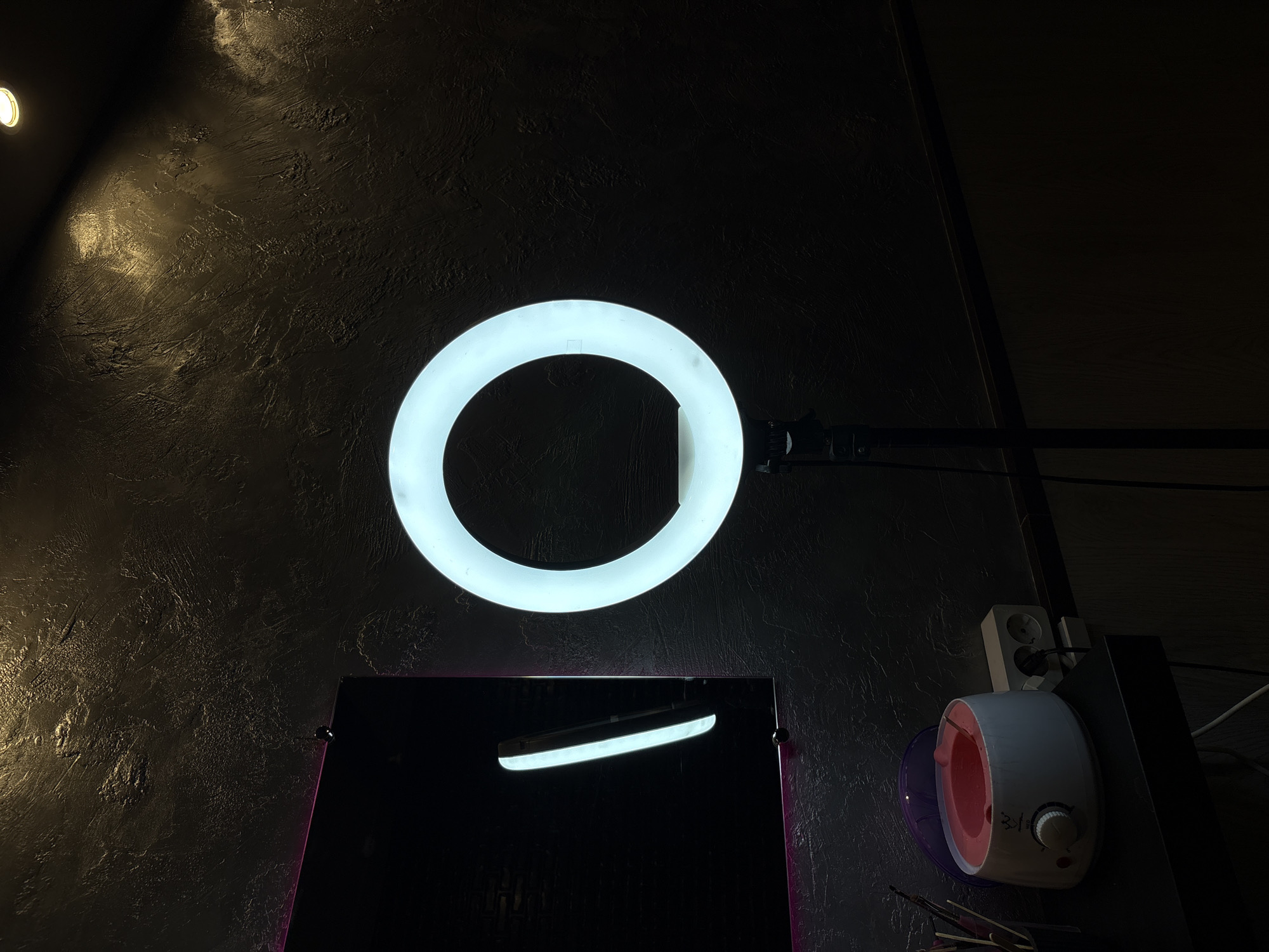 The image depicts an indoor scene, possibly a salon or a personal beauty space, with a dark, textured wall as the backdrop. A prominent ring light, illuminated with a bright white glow, is centrally positioned on a stand. To the left of the ring light, a mirror with a subtle pink LED trim is visible, reflecting a vertical light strip. Below the mirror and to the left of the ring light, on a black shelf, is a pink wax warmer with its lid open, and a dial on its front. A multi-socket power strip is plugged into the wall behind the wax warmer. The lighting in the scene is dim, with the ring light and a ceiling light providing illumination. There are no people visible in the image. The specific location in Rostov-on-Don, Russia, cannot be determined from the visual information provided in the image.