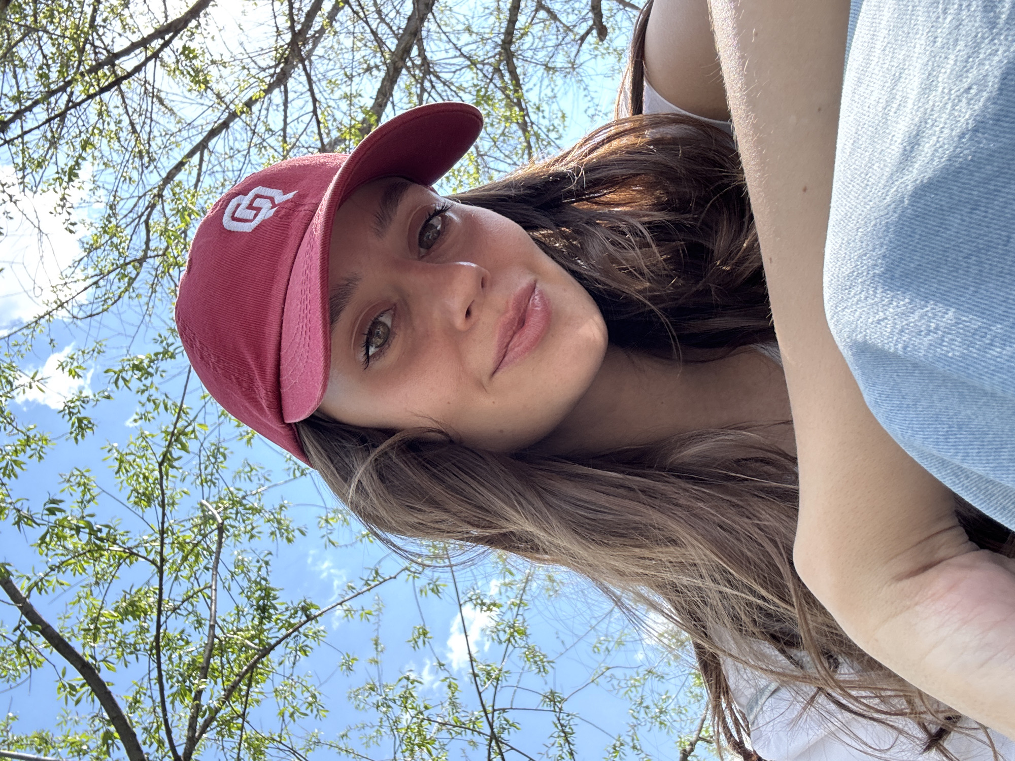 A young woman with fair skin and brown hair is facing the camera. She is wearing a red baseball cap with a white logo on the front. She is wearing a white top and a light blue denim jacket or shirt. The background shows a blue sky with tree branches and young green leaves. The location of the city of Овсянка, Russia cannot be confirmed from the available image.