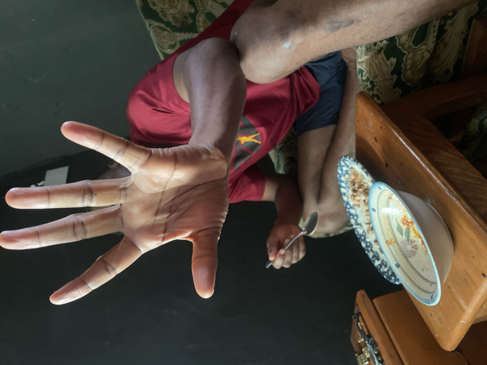 A person's hand, fingers spread wide, is in the foreground, obscuring the face of a person in the background. The person in the background is wearing a red t-shirt and dark shorts and is sitting on a couch with a patterned upholstery. They are holding a spoon and appear to be eating from a plate of food. In front of the couch, there is a wooden table with a bowl of food and another plate of food. To the left of the table is a wooden chest. The scene is dimly lit and appears to be indoors. This image was taken in Jos, Nigeria.