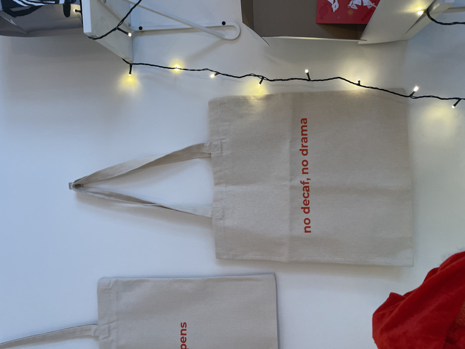 The image displays an interior scene, likely a room or retail space, featuring a white wall. Two beige canvas tote bags are visible. The central tote bag is suspended from a small, silver-colored hook on the wall, displaying the red text "no decaf, no drama." To its left, a second, partially visible beige tote bag also hangs on the wall, showing the red text fragment "pens" in its upper portion. On the right side of the frame, a string of warm white LED fairy lights hangs vertically, casting a soft glow. These lights are draped over and beside a white shelf unit. On the shelf, a white box is partially open, revealing a red item inside, possibly a greeting card or gift wrap with white designs. Above this shelf, a black and white striped object, possibly a bag or footwear, is partially visible. In the bottom left corner of the image, a crumpled red fabric item is present. The specific location within the city of Belgrade, Serbia, cannot be determined from the visual evidence provided.