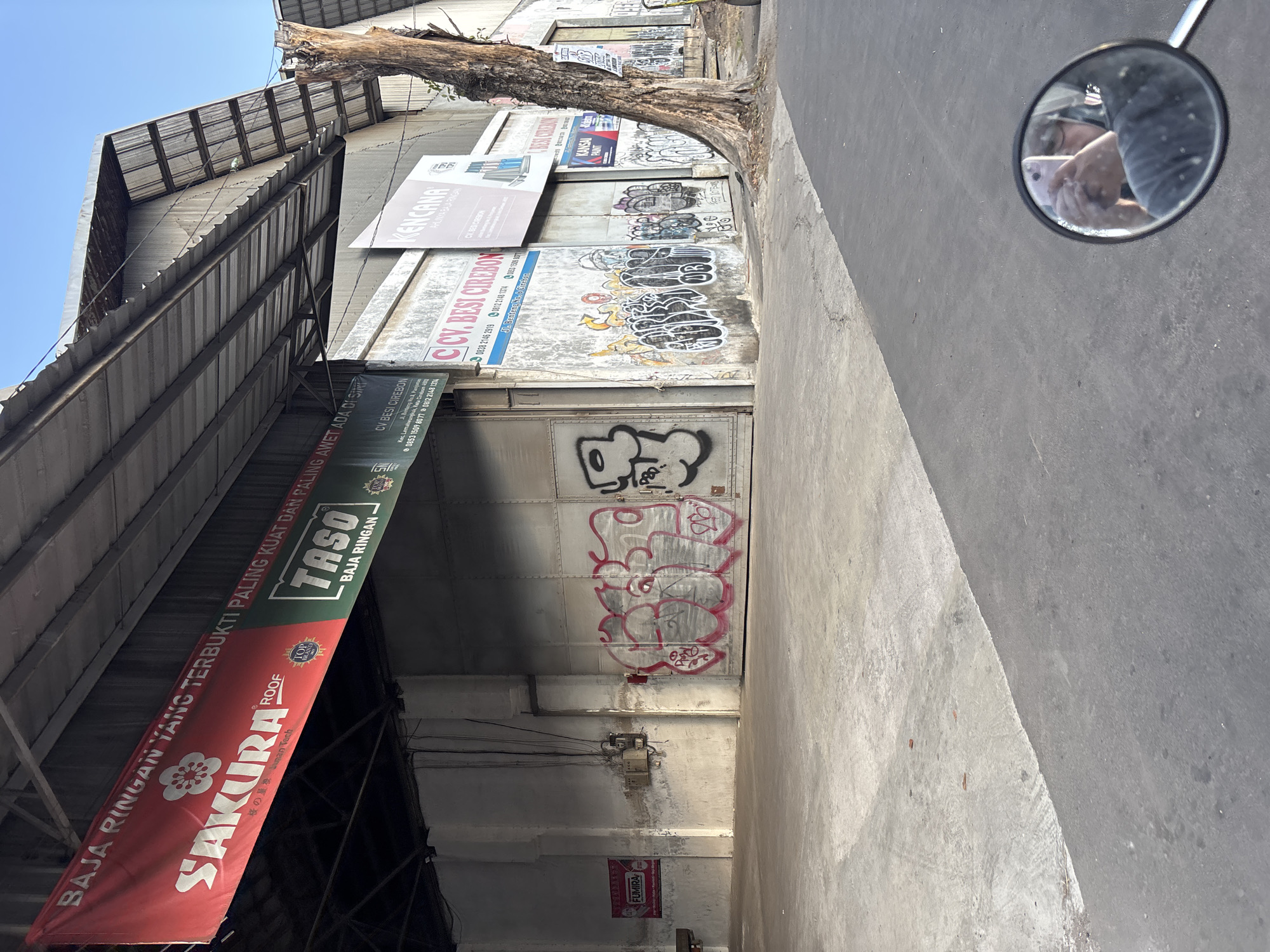 The image captures a street view in Cirebon, Indonesia, focusing on the exterior of a commercial building. The building features a large, metal roller door, much of which is covered in graffiti. The facade is also adorned with several banners and signs. One prominent red banner displays the "SAKURA ROOF" logo, alongside text in Indonesian. To its right, a green banner advertises "TASO BAJA RINGAN". Further along the building, there are signs for "CV. BESI CIREBON", "KENCANA", and "KANSAI PAINT". The ground in front of the building is paved with concrete, showing some wear and tear. A large tree with a thick trunk stands near the right edge of the frame, its bark weathered and textured. In the foreground, the reflection in a motorcycle's side mirror shows a person holding a phone and taking a selfie, indicating the perspective of someone on a motorcycle passing by. The sky above is clear and blue. The overall impression is one of an industrial or commercial area in an urban setting, with signs of business activity and street art.