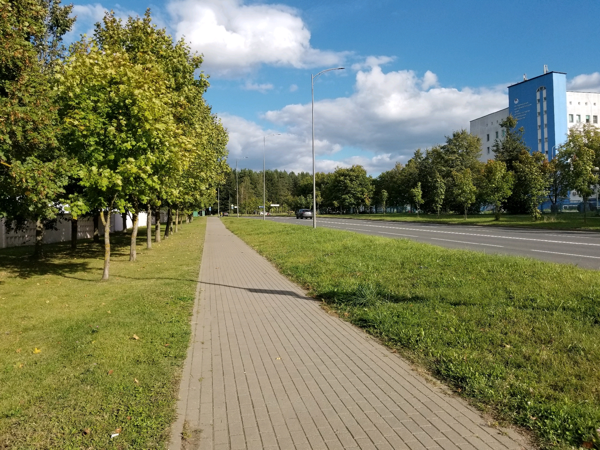 The image shows a paved sidewalk running alongside a grassy area and a road with multiple lanes. Trees line the sidewalk on the left, and a building with blue and white sections is visible in the background on the right. Several streetlights are present along the sidewalk. The sky is blue with scattered white clouds. The location is identified as Бараўлянскі сельскі Савет, Belarus. A dark-colored car is driving on the road in the distance. The overall scene depicts a well-maintained urban or suburban street with pedestrian and vehicular infrastructure.