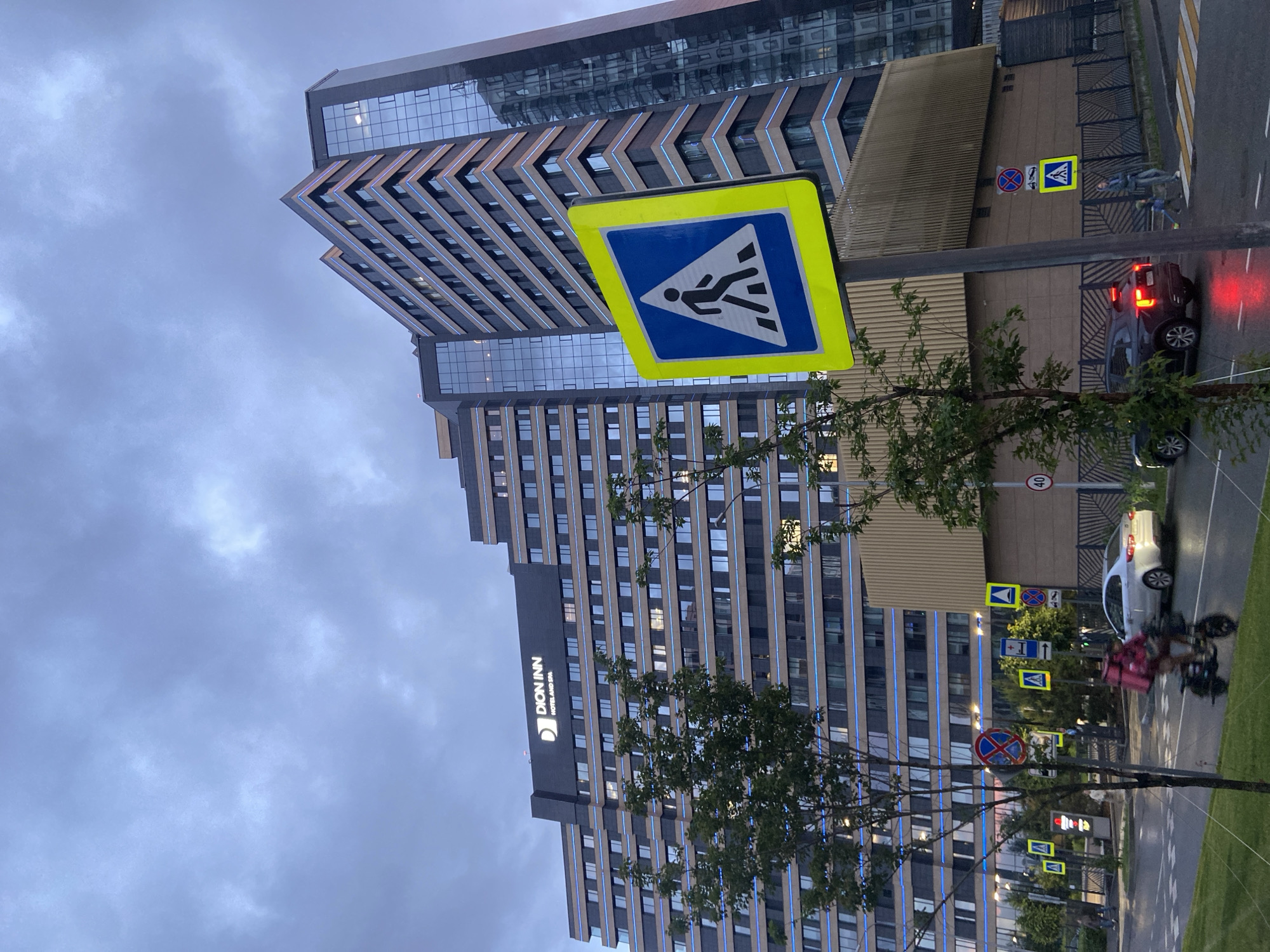 The media file captures an urban scene in Kazan, Russia, during what appears to be a heavily overcast day or early dusk, given the muted light and illuminated building accents. The image is oriented vertically (rotated 90 degrees clockwise from its natural upright position).
In the foreground, a prominent yellow-bordered blue sign for a pedestrian crossing stands on a pole, slightly left of the center. Behind it, a modern, multi-story building dominates the left side of the frame. This building, likely a hotel, features a sleek design with large glass windows and horizontal bands, some of which exhibit a distinctive blue lighting or detailing. The text "DION INN HOTEL AND SPA" is visible as illuminated signage on its facade.
To the right of the foreground sign and further down the street, another contemporary building with a similar stepped or tiered design and glass facade is visible. The scene includes a paved road with a zebra crossing. A dark-colored SUV or car with red taillights is seen on the road, possibly stopped or moving slowly. Further along the street, a white car is also visible.
Several traffic signs are scattered along the roadside, including another pedestrian crossing sign, a "No Stopping" sign (blue circle with red border and diagonal line), and a circular speed limit sign showing "40". On the right side of the street, near a crosswalk, two individuals are present: one on a kick scooter and another person walking. Further down, another person on a scooter or bicycle, wearing a prominent pink or red delivery backpack, is also visible. Green trees with lush foliage are interspersed throughout the scene, partially obscuring the buildings and traffic elements. The sky is completely overcast with light grey clouds.