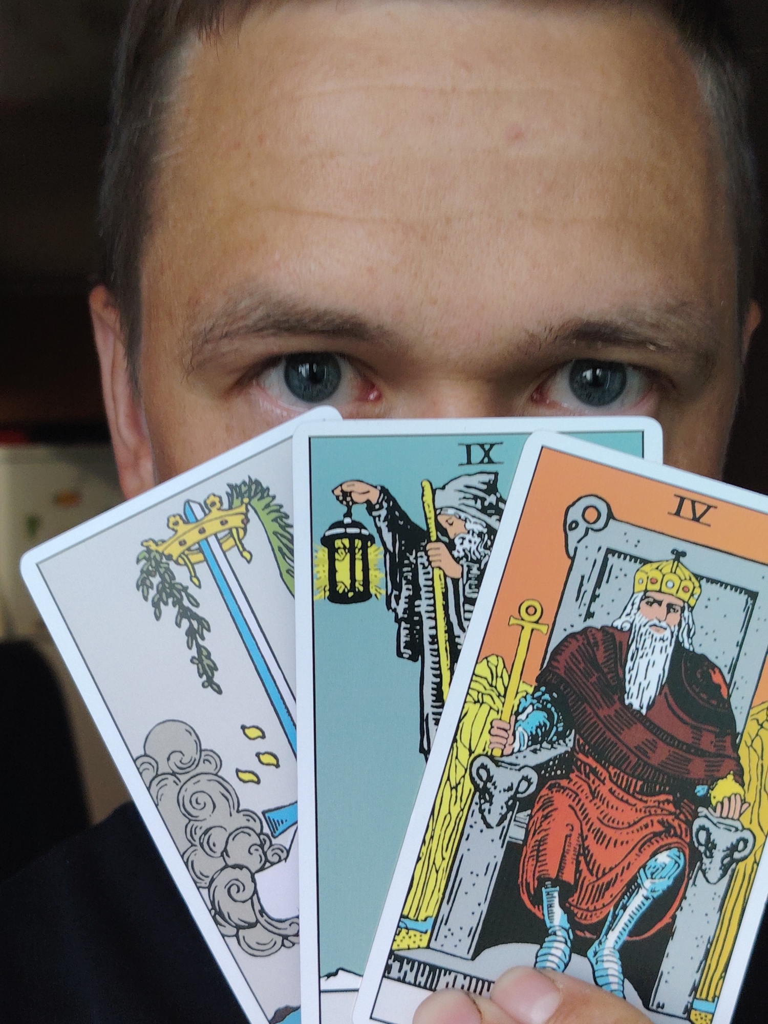 The image shows a close-up of a person's eyes and forehead, looking directly at the camera. The person is holding three tarot cards in front of their face, obscuring the lower part of their face. The cards visible are the "IX of Wands," a card featuring a figure holding a lantern, and the "IV of Wands," which depicts a bearded king seated on a throne. The background is dark and out of focus, suggesting an indoor setting. There is no identifiable information about the location within the city of Chaikovsky, Russia, as the scene is entirely focused on the person and the tarot cards.