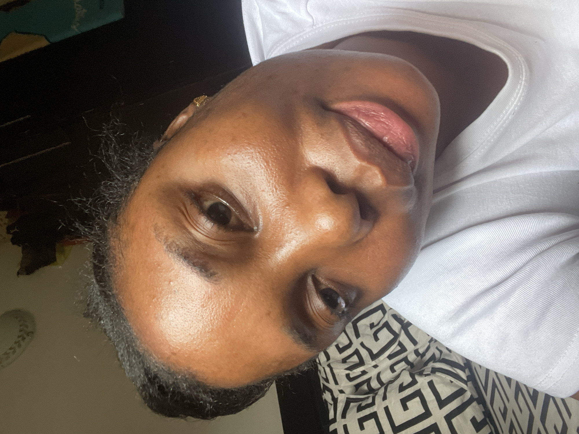 Close-up view of a dark-skinned woman's face with her head tilted to the right, she has dark hair, her face is shiny, wearing a white shirt, and a small gold earring in her left ear, lying on a white and black patterned fabric, location unknown, presumably indoors.
