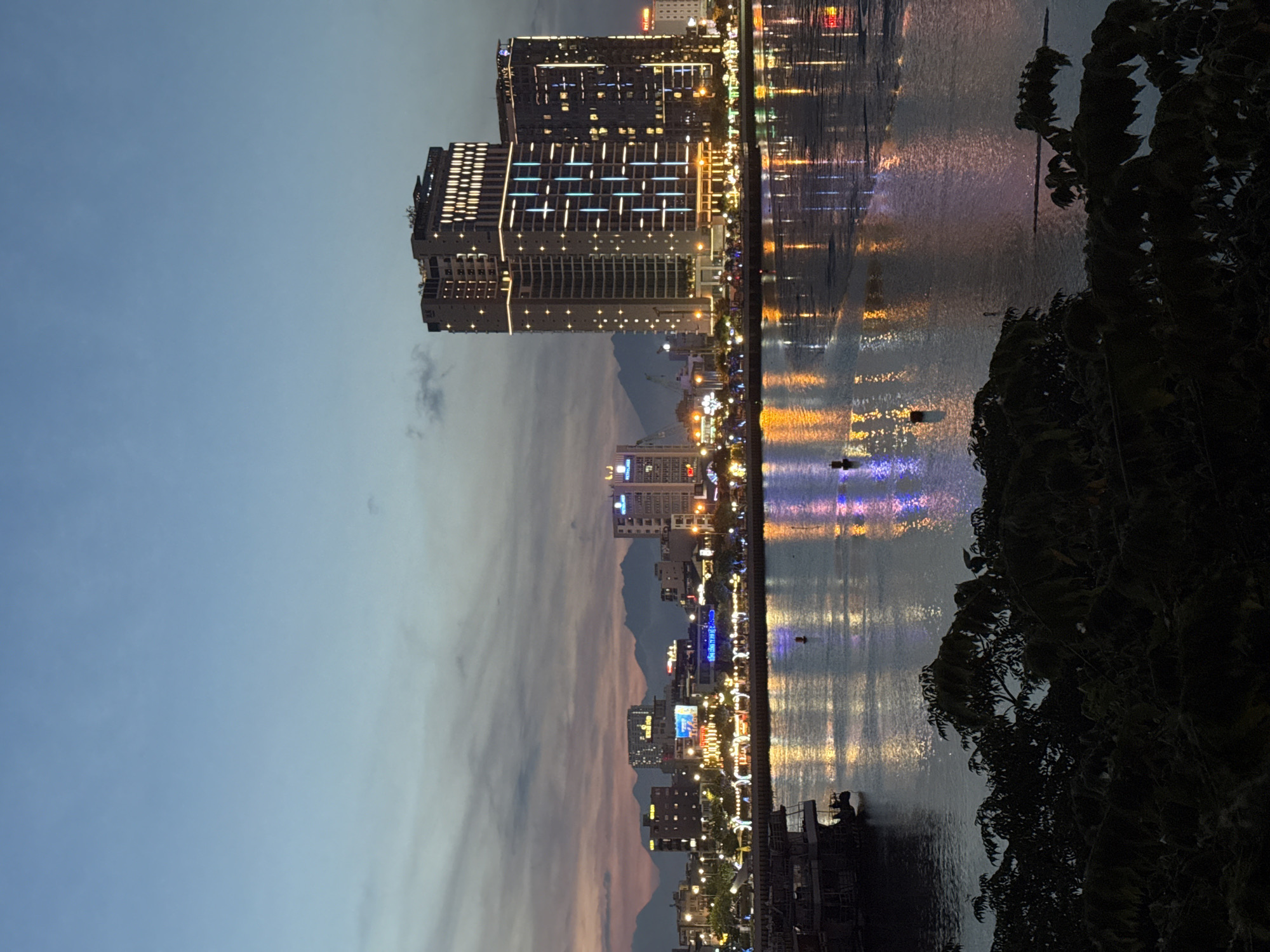 The image depicts a twilight scene of a city skyline along a waterfront in Da Nang, Vietnam. Tall buildings, illuminated with numerous lights, line the shore. The lights from the buildings are reflected on the calm surface of the water, creating shimmering patterns of yellow, blue, and purple. A silhouette of dark foliage frames the right side of the image, adding depth to the scene.
The sky is a gradient of soft blues and muted pinks, characteristic of dusk. Distant mountains are faintly visible behind the city, shrouded in the dim light. A few small boats or structures are present in the water, marked by distant lights. There appears to be a walkway or promenade along the waterfront, with more lights suggesting a populated area. One person is visible as a distant silhouette in the water.
The overall mood is serene and peaceful, capturing the transition from day to night in a vibrant urban environment. The time of day is clearly evening or twilight, with artificial lights beginning to dominate the scene. No legible text is visible in the image.