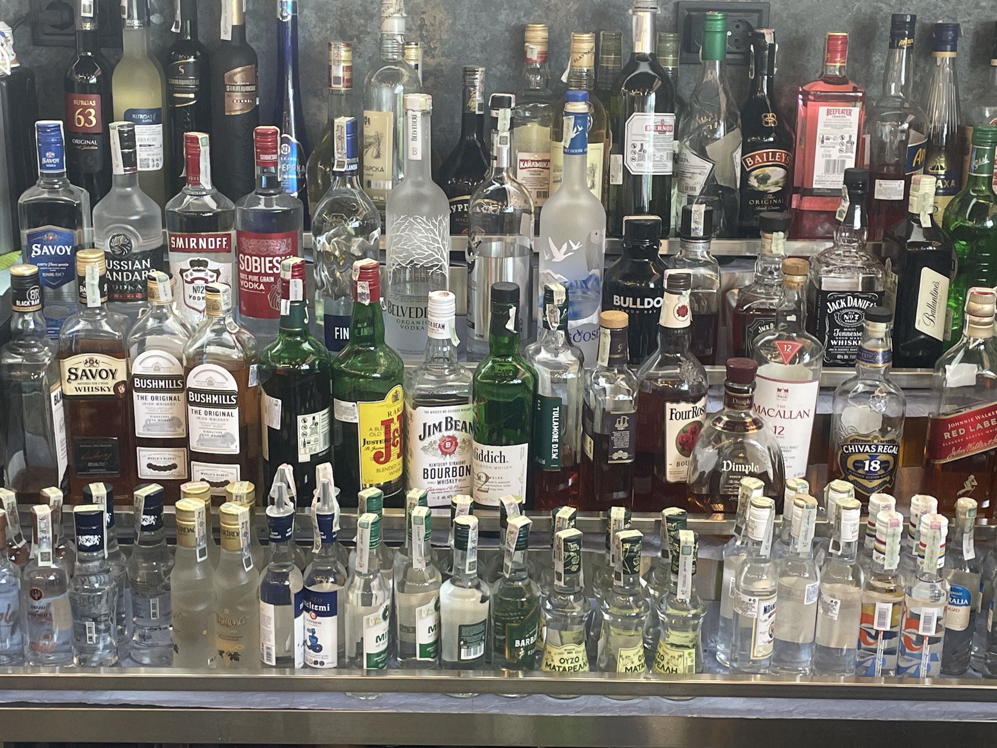 The image depicts a well-stocked bar with multiple shelves displaying a variety of alcoholic beverages. The bottles include vodkas (such as Savoy, Russian Standard, Smirnoff, Sobieski, Belvedere, Grey Goose, and Karawanska), whiskies (such as Bushmills, Jim Beam, Four Roses, Macallan, Chivas Regal, and Johnnie Walker), and other spirits like Baileys, Beefeater, and Ouzo. Many of the bottles on the lowest shelf are smaller, possibly miniature, sizes. The bar setting appears to be indoors.