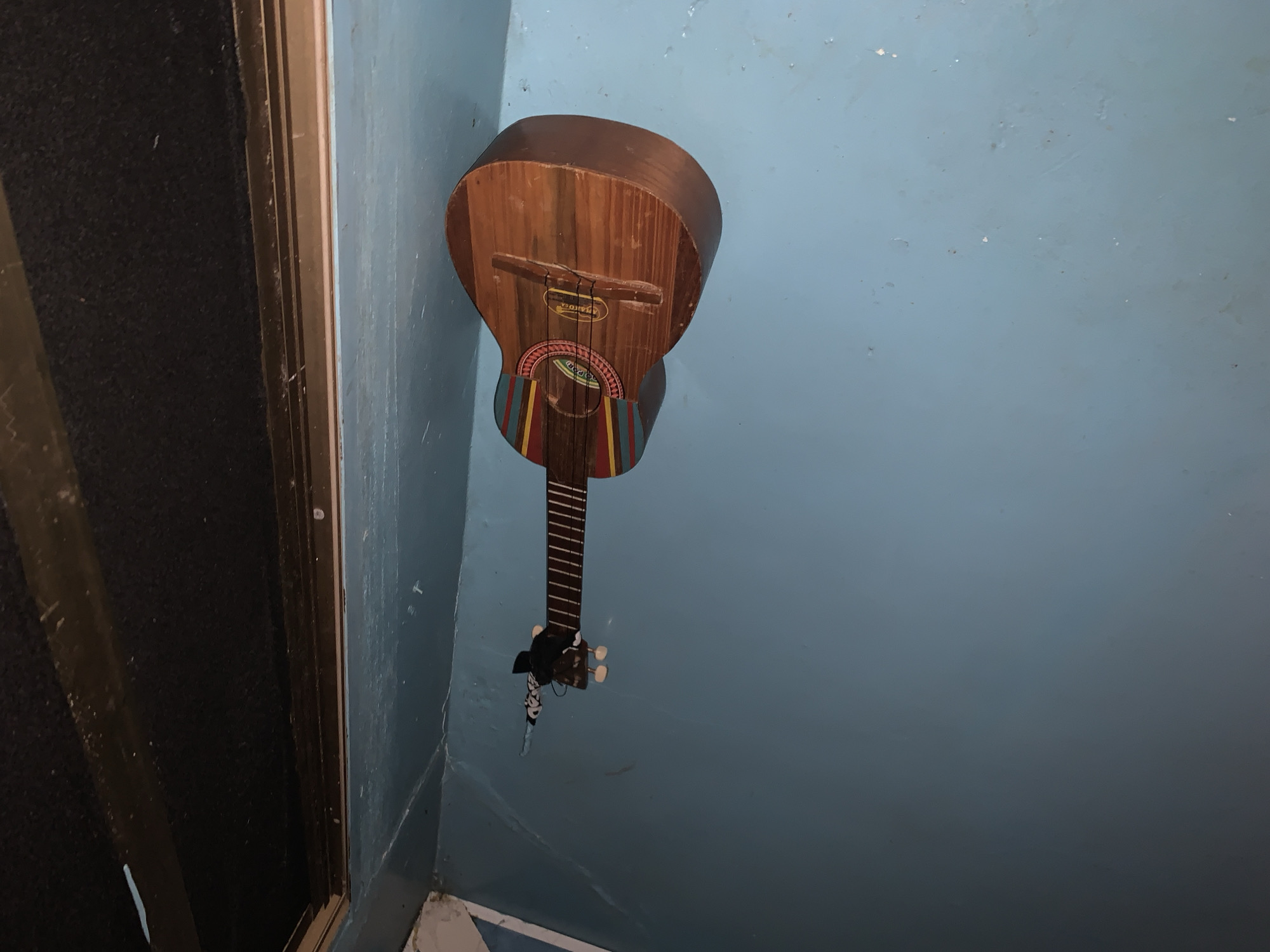 The image displays an indoor scene, likely in Santa Ana, Venezuela, centered on a wooden string instrument leaning against a textured, light blue painted wall. The instrument, resembling a cuatro or small guitar, features a natural wood finish. Its circular sound hole is adorned with a vibrant, multi-colored geometric design of red, yellow, blue, and green stripes. Near the headstock, a piece of black and white patterned fabric is tied to the instrument's neck, which has dark frets and light-colored tuning pegs.
Visible on the instrument's soundboard, above the sound hole, is text including "VENEZUELA" and another partially obscured word, possibly "CORAL". To the left of the instrument, a dark, textured vertical structure with a metallic frame runs along the edge of the scene; this could be a door, a window with a curtain, or a large speaker. The blue wall exhibits some imperfections and texture. The lighting is subdued, suggesting an interior space without bright direct light. No activity is depicted in the static scene.