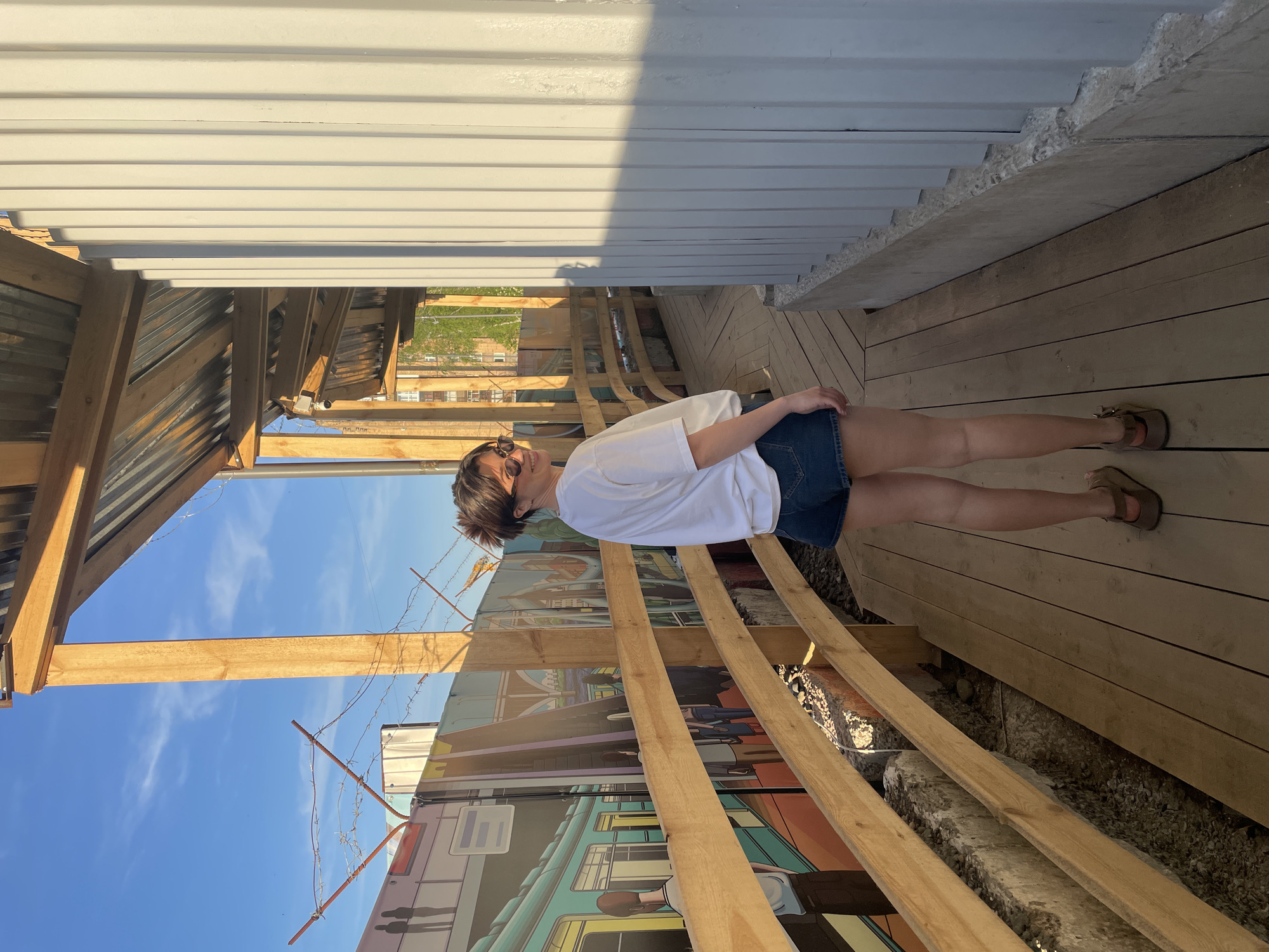 A young woman stands outdoors on a sunny day in what appears to be an urban or semi-urban setting in Krasnoyarsk, Russia. She is positioned on a wooden deck or boardwalk, facing slightly away from the camera and looking towards the left side of the frame, with a relaxed expression and a subtle smile. She has short, dark hair and wears sunglasses pushed up on her head. Her attire consists of a white t-shirt, blue denim shorts, and open-toed sandals.
To her right, a tall, white corrugated metal wall or building facade casts a shadow, indicating bright sunshine. To her left and behind the wooden railings, a vibrant, colorful mural depicting buildings, possibly a cityscape, adorns a wall. Above the mural, barbed wire is strung across wooden posts. The sky above is clear and blue with some scattered white clouds. To the far left, a structure with a corrugated metal roof and wooden beams is visible. Below the wooden decking, gravel and concrete elements are present. The overall scene suggests a casual outdoor environment, possibly a pedestrian area, a patio, or an artistic space, enjoying pleasant weather. No discernible text is visible in the image.