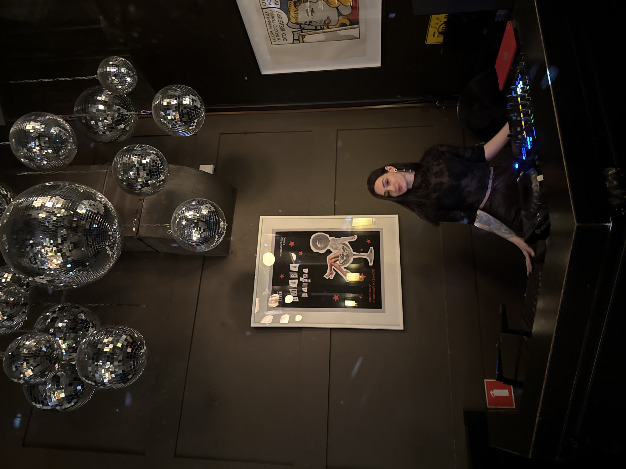 The image shows a DJ booth in what appears to be a bar or club, possibly located in Moscow, Russia, given the Cyrillic script on a comic-style framed print on the wall. A woman with long dark hair and a lace top is standing at a DJ console with illuminated controls. Next to the DJ booth, a dark wall serves as a backdrop for several shiny disco balls, and framed posters, one appearing to be a collage featuring a dancer figure, another displaying Russian text with comic book graphics. A small red fire extinguisher sign is affixed to the wall near the right corner.