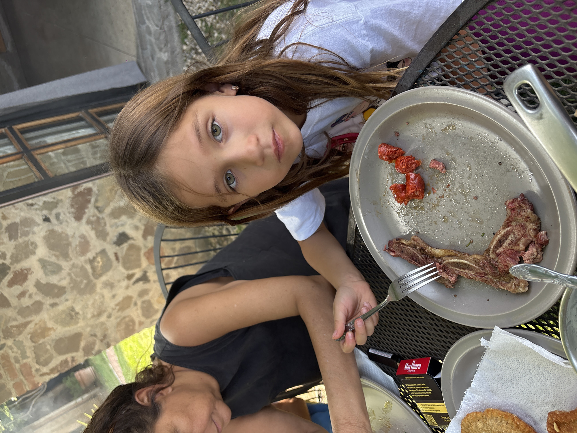 A girl with long brown hair is looking up at the camera next to a plate with meat and sausages on a metal table. A woman with dark hair is holding a fork and cutting the meat. A pack of Marlboro cigarettes is on the table, next to a plate with breaded food. The setting appears to be outdoors, possibly a patio, in San Bernardino Tlaxcalancingo, Mexico.