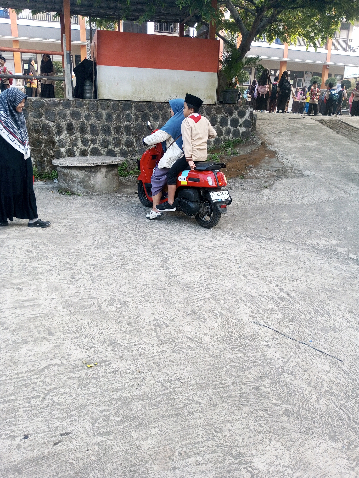In front of a red and white building facade, two individuals are seated on a red scooter. The person in front wears a blue headscarf, while the person behind wears a light brown jacket and a black peci hat. Both are looking away from the camera. The scooter is parked on a grey concrete surface. To the left, a person in a black hijab and skirt stands observing the scene. In the background, other individuals, many appearing to be students wearing uniforms and carrying backpacks, are gathered near the building. The environment appears to be an outdoor area, possibly a schoolyard, with a large tree providing shade and a stone wall in the mid-ground. The license plate of the scooter reads "AA 6515 TZ". The location is identified as Krasak, Indonesia.